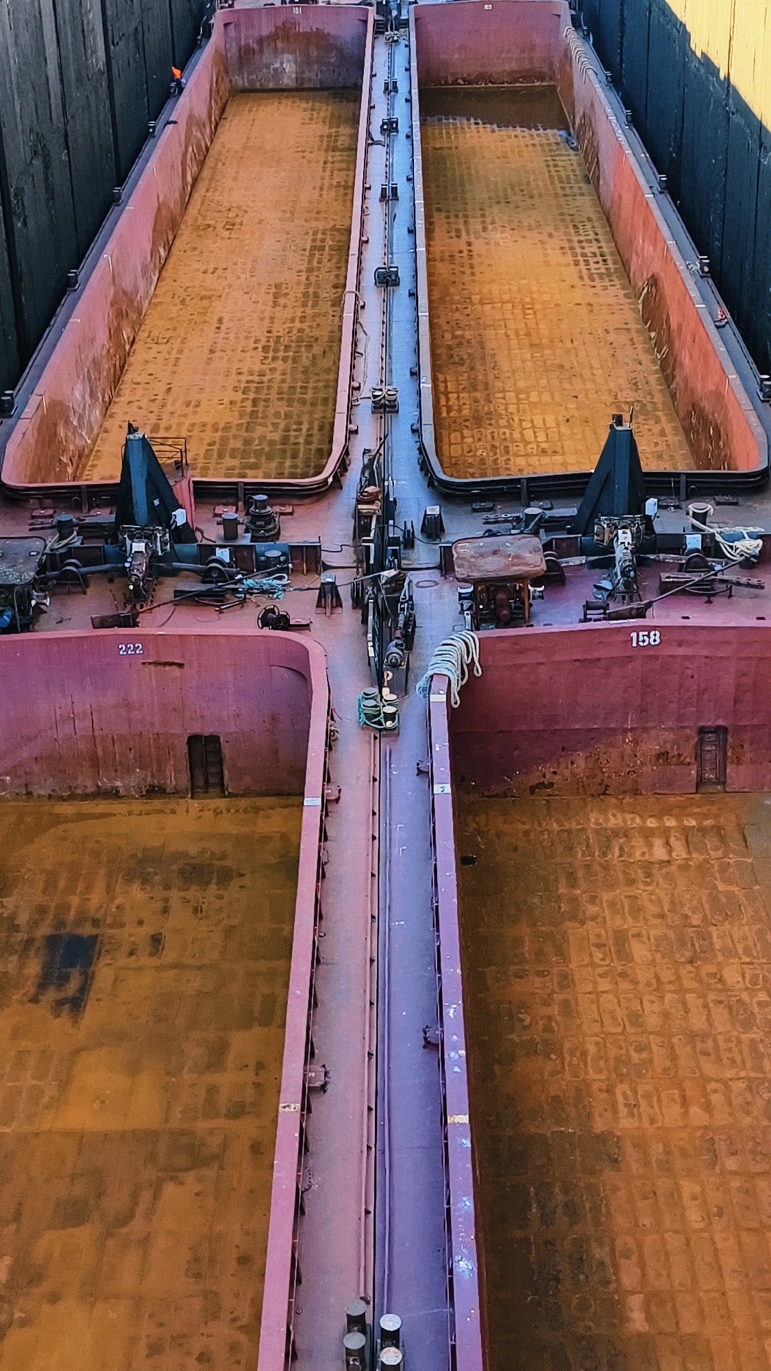The image shows an aerial view of two large, empty barge holds, likely at a port or industrial facility. The holds are reddish-brown and appear to be constructed of metal. They are separated by a central walkway or structure. There are numbers visible on the sides of the barges, "222" and "158". Various pieces of industrial equipment, including winches and ropes, are visible on the decks. The floor of the holds has a textured pattern, suggesting it is designed to carry cargo. The surrounding environment appears to be industrial, with a dark wall or structure visible on the left side of the image. The scene is bathed in sunlight, casting shadows and highlighting the textures of the barge.