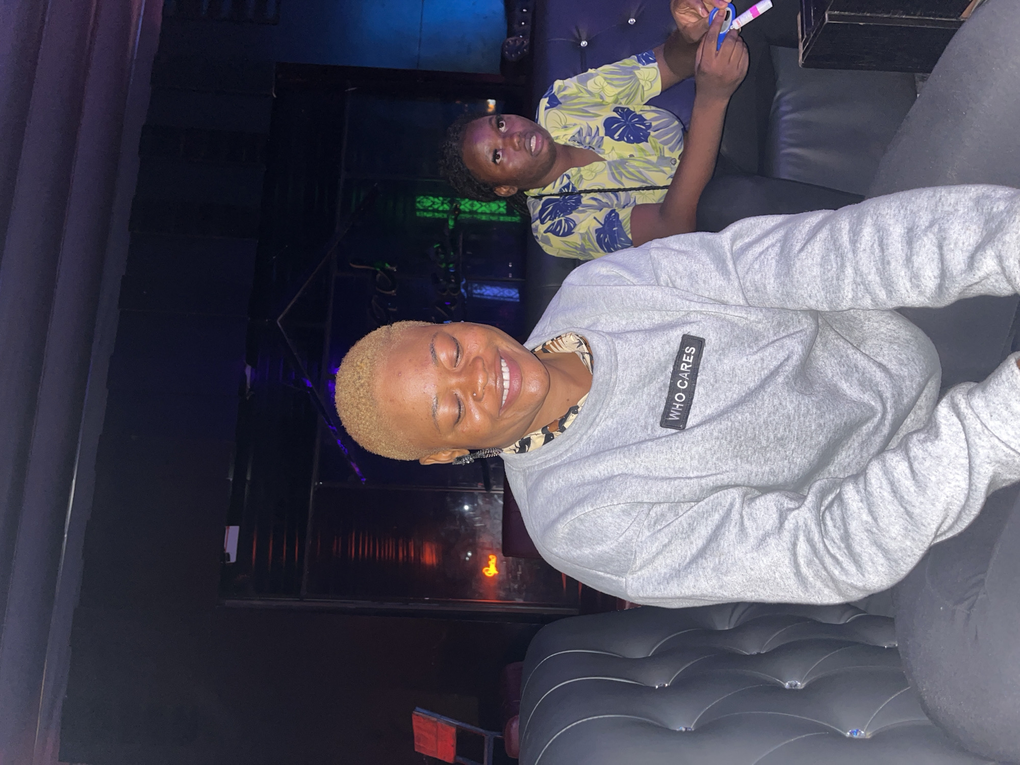 This image depicts an indoor scene within a dimly lit establishment, resembling a lounge or bar, located in Howayi, Nigeria. Two females are seated on a dark, button-tufted leather sofa or booth.
In the foreground on the left, a female with short, bleached blonde hair and a nose ring on her left nostril is positioned, facing slightly to her right. Her eyes are closed, and she is smiling broadly, revealing her upper teeth. She wears a grey long-sleeved crew-neck sweatshirt with a black rectangular patch on the upper chest that reads "WHO CARES" in white text. A patterned, collared shirt is visible beneath her sweatshirt at the neckline. Her lower body is clad in dark-colored pants.
In the background on the right, another female with dark, braided hair is seated, looking upwards and to her right with wide eyes and an open mouth. She wears a short-sleeved button-up shirt featuring a light yellow background with a blue and green tropical leaf or floral pattern. In her right hand, she holds a small, blue-handled pair of scissors and a white and pink cylindrical object, possibly a pen or marker.
The background consists of dark walls with various artificial light sources. To the left behind the foreground subject, a red chair or object with a light frame is partially visible. Further back, an orange-red neon-style light in the shape of a partial letter (possibly 'D' or 'O') is visible. Green and blue vertical light elements are also present. The ceiling above appears dark and potentially paneled. To the far right, a dark, rectangular object, possibly a table or box, is partially visible next to the sofa.