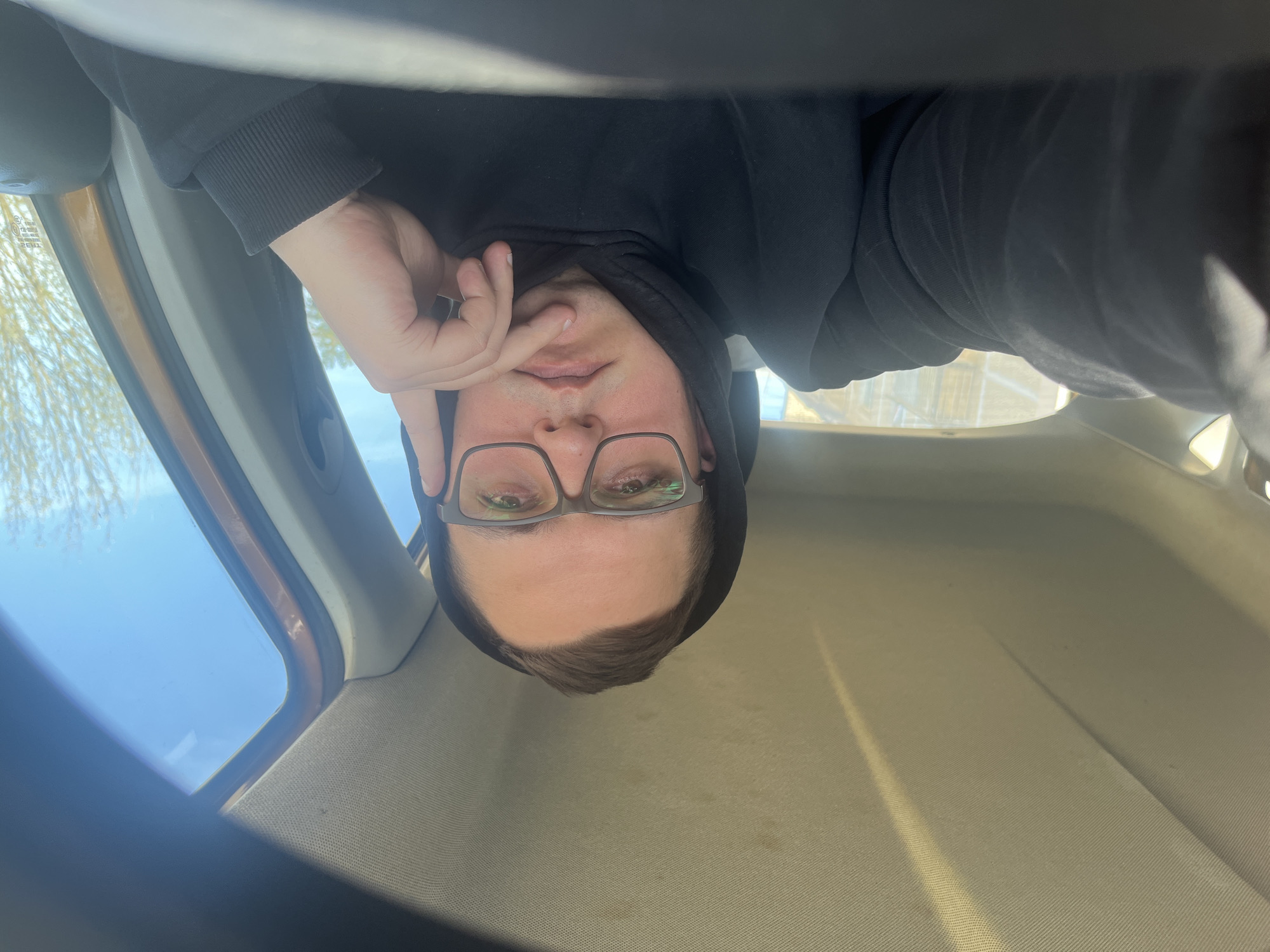 The media file depicts a person taking an upside-down selfie inside a car. The individual, appearing to be male, is wearing a black hooded sweatshirt and glasses, looking directly at the camera with a neutral expression. A hand is visible near their face, with fingers touching the chin area. The setting is the interior of a vehicle, specifically a car, with a light-colored headliner and a window frame visible. Outside the window, a clear blue sky is visible, indicating bright daylight and sunny weather. Mirrored text "TRB2", likely a vehicle marking, is visible on the car window. The location is Razdolye, Russia.