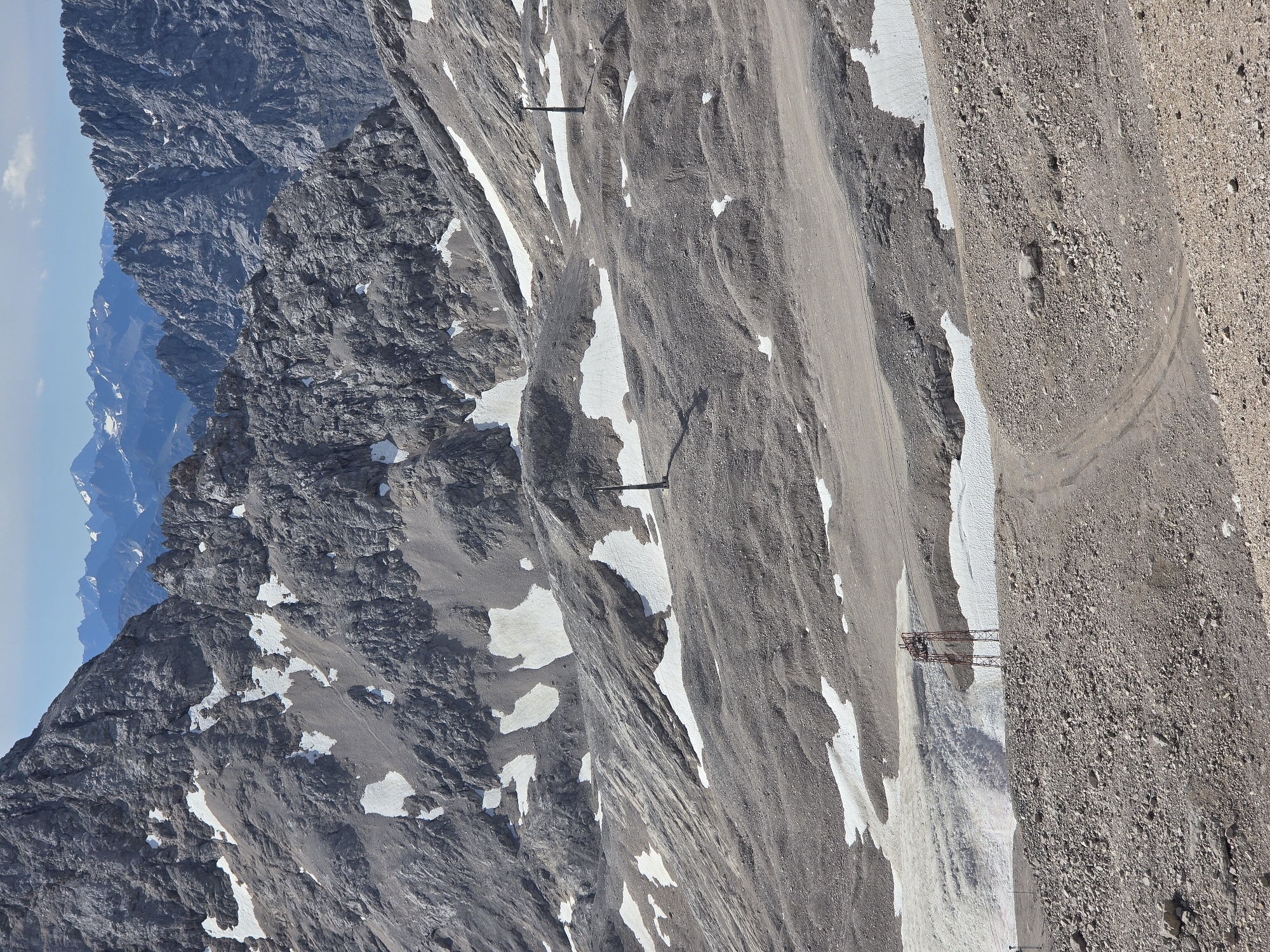 The image depicts a rugged, rocky mountain landscape with patches of snow. In the foreground, a dirt path winds through gravel and scattered rocks. Further back, several dark poles are visible, likely part of a ski lift or similar infrastructure. One prominent structure in the mid-ground is a rusty, lattice tower with a cable extending from it. The terrain is characterized by undulating slopes, steep rock faces, and areas of debris. The background reveals a range of distant, sharp mountain peaks under a clear blue sky with a few scattered clouds. This landscape is consistent with the high alpine environment of the Zugspitze, Germany's highest peak, located near Garmisch-Partenkirchen. No people are visible in the image.
