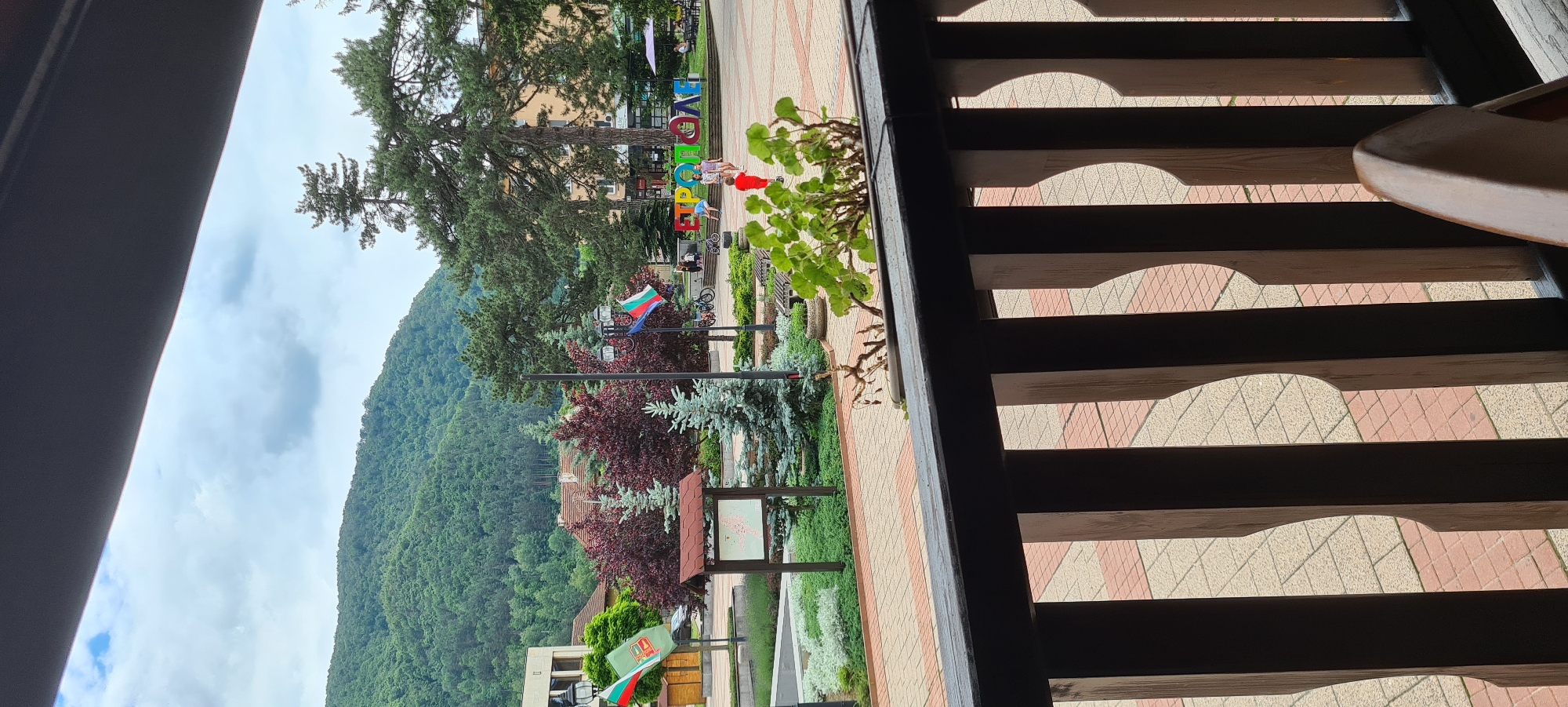 The image presents an outdoor scene viewed from a raised perspective, likely a balcony or terrace, in the city of Etropole, Bulgaria. In the immediate foreground, a dark wooden railing with decorative vertical slats partially obstructs the view. To the right, a light-colored chair arm is visible. Directly beyond the railing, a potted plant with vibrant green leaves stands, while another dried plant is also present.
The midground reveals a paved town square or park area, consisting of light-colored and reddish interlocking tiles. The area is landscaped with various types of greenery, including low-lying shrubs, several small blue spruce-like conifers, and larger deciduous trees with reddish foliage, as well as tall evergreen trees like pines. Several streetlights are visible, along with wooden benches. A brown information board or map stand is positioned near the center-left. Two prominent flagpoles display Bulgarian flags (white, green, red horizontal stripes), and one pole also shows a blue flag with yellow stars, likely representing the European Union.
People are scattered across the square: a few children, one notably in a bright red shirt, are walking or playing. Some adults are also present, and bicycles are parked nearby. The most striking feature in the midground is a large, colorful, three-dimensional sign spelling out "ЕТРОПОЛЕ" (Etropole) in Cyrillic script.
The background features multi-story buildings, some with visible shops on their ground floors, indicating commercial activity. Behind the urban structures, a range of densely forested green mountains rises prominently under a partially cloudy blue sky, forming a natural backdrop to the town.