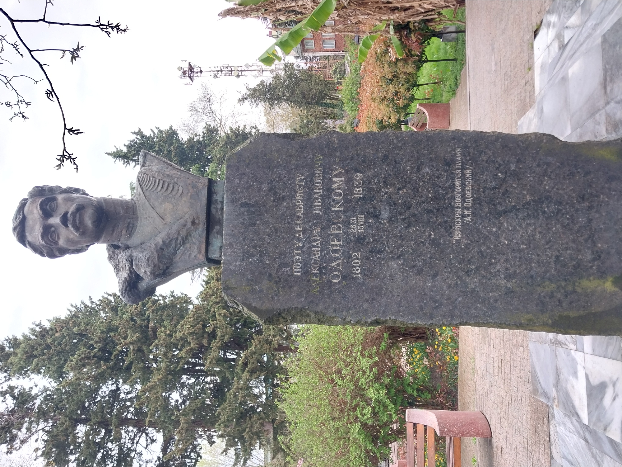 The image shows a bust monument of a man with his name engraved on a dark gray stone slab. The inscription indicates that this monument is dedicated to the poet and Decembrist Aleksandr Ivanovich Odoyevsky (1802-1839). The Russian inscription includes a quote attributed to Odoyevsky: "From sparks, a flame will ignite / A. I. Odoyevsky."
In the background, there's greenery including bushes, trees, and potentially a banana tree. A communications tower is visible in the distance. The monument is situated on a paved area next to a walkway and a bench. The photograph was taken in Sochi, Russia.