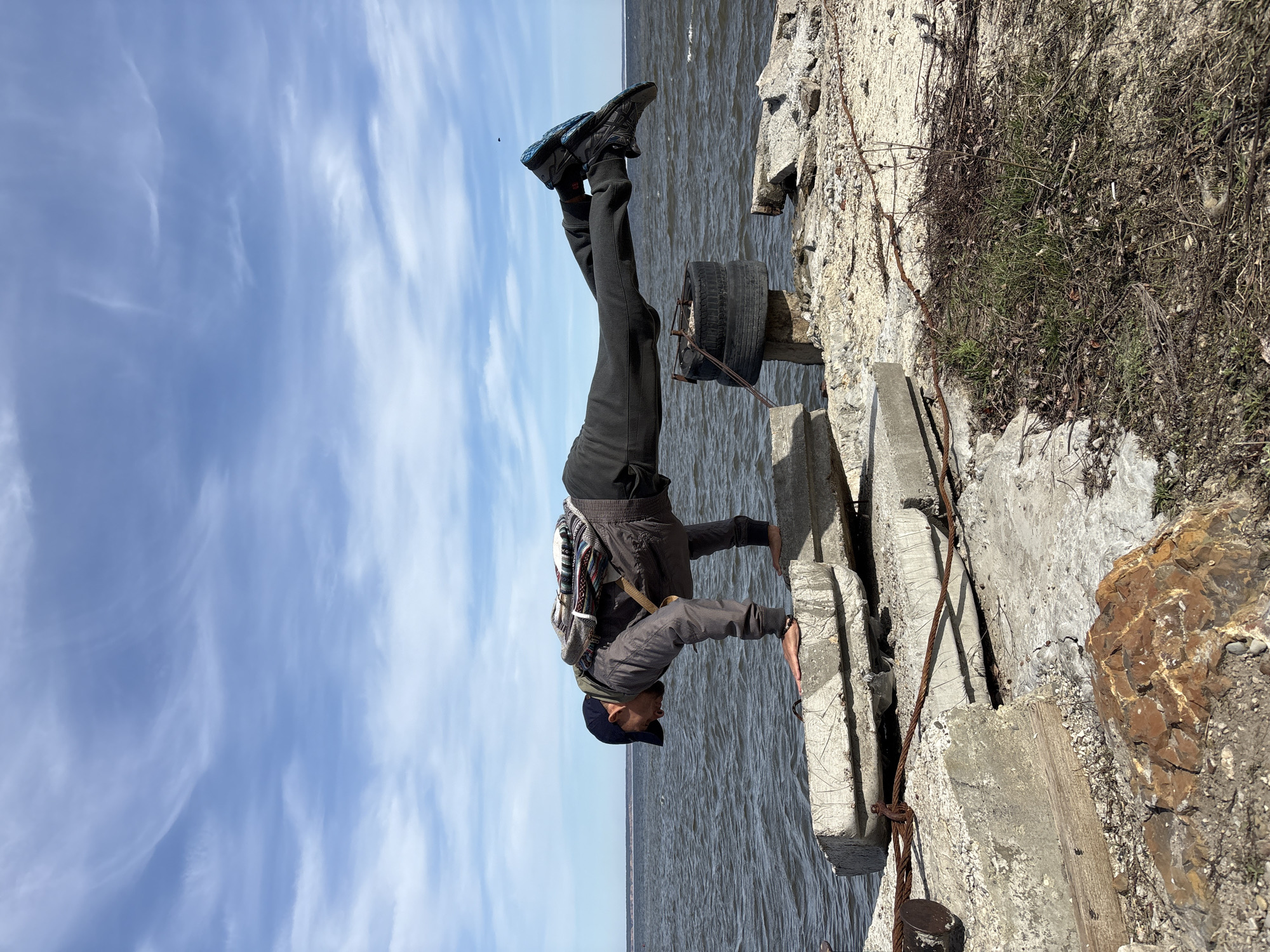 The media file captures an outdoor scene featuring a person performing a handstand on a concrete structure next to a body of water. The image itself is rotated 90 degrees counter-clockwise.
The primary subject is an adult, likely male, dressed in dark pants, a dark jacket, and a dark baseball cap. He wears a backpack with a multi-colored, possibly patterned, design. He is inverted, supporting himself on his hands, which are placed on weathered concrete blocks. His legs are extended upwards, and he is wearing athletic shoes.
The setting is an embankment or pier beside a large body of water, which extends to the horizon. The water surface shows ripples, suggesting a gentle breeze or current. Above the horizon, a bright blue sky is visible, adorned with scattered, wispy white clouds. The immediate foreground and mid-ground consist of rough, aged concrete blocks and slabs, with patches of dry grass, weeds, and some bare soil. A discarded, dark-colored tire is resting on one of the concrete structures, and rusty metal elements, possibly rebar or cables, are integrated into the concrete.
The activity taking place is a display of physical strength and balance, with the person executing a handstand. The time of day appears to be bright daylight, likely late morning or early afternoon, given the clear conditions. The location details suggest a waterfront area in Ulyanovsk, Russia, possibly along the Volga River. There is no visible text in the image.