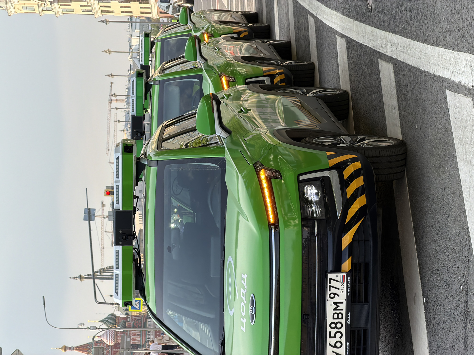 A row of green-colored, autonomous vehicles with roof-mounted sensor arrays are parked on a city street near Red Square in Moscow, Russia. The vehicles feature yellow and black hazard striping on their front corners and Cyrillic script logos on their hoods. A Russian license plate is visible on the front of the closest vehicle. A crosswalk is evident beneath the parked vehicles. In the background, the distinctive architecture of Moscow, including the domes of St. Basil's Cathedral and other buildings, can be seen, along with construction cranes. People are visible in the lower left of the frame. The sky is overcast.