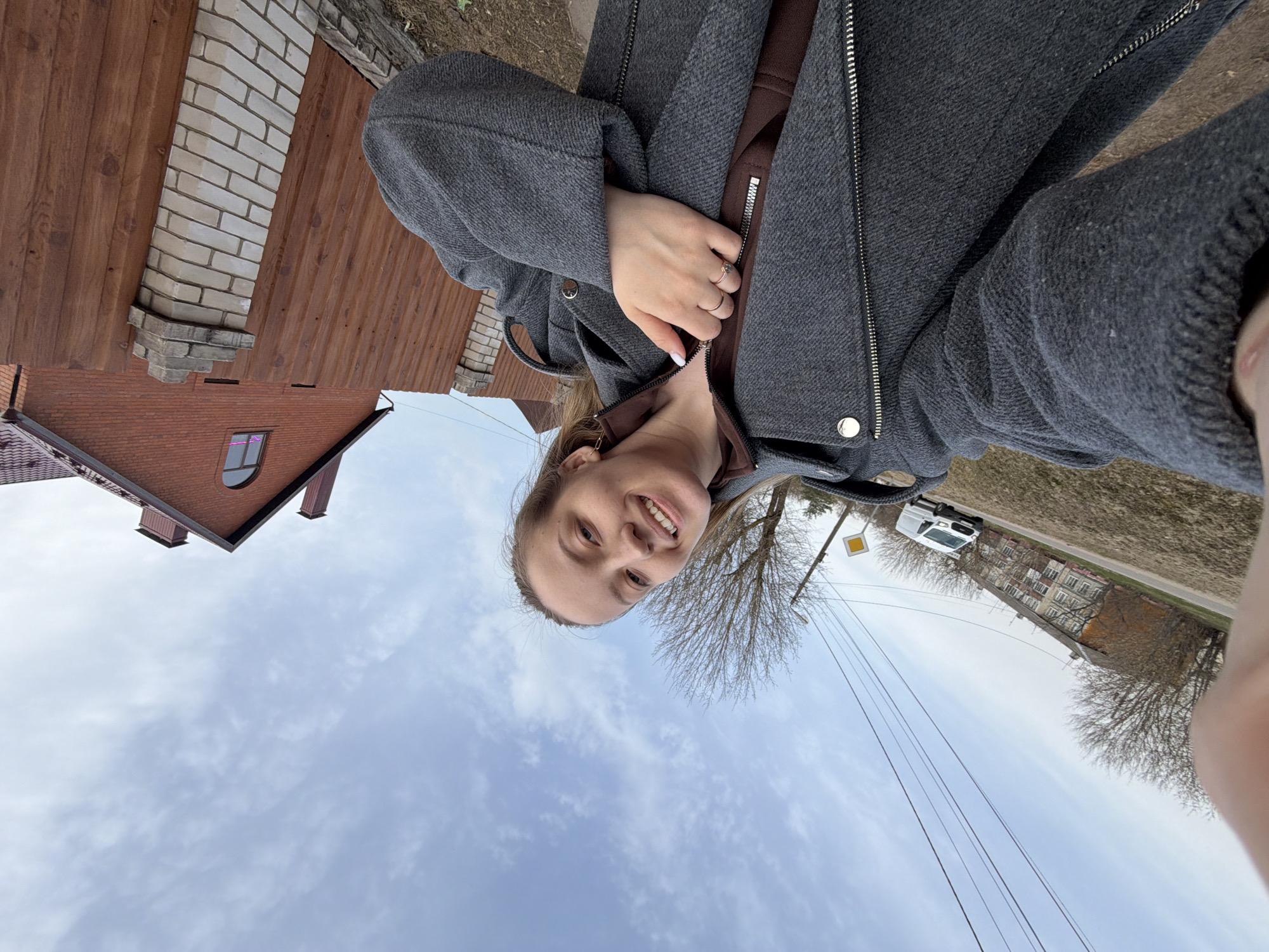 A woman in a gray jacket poses for a selfie. In the background are houses, a van, a power pole with wires, and a tree without leaves. The sky is cloudy. The photo may have been taken in Reshetnikovo, Russia.