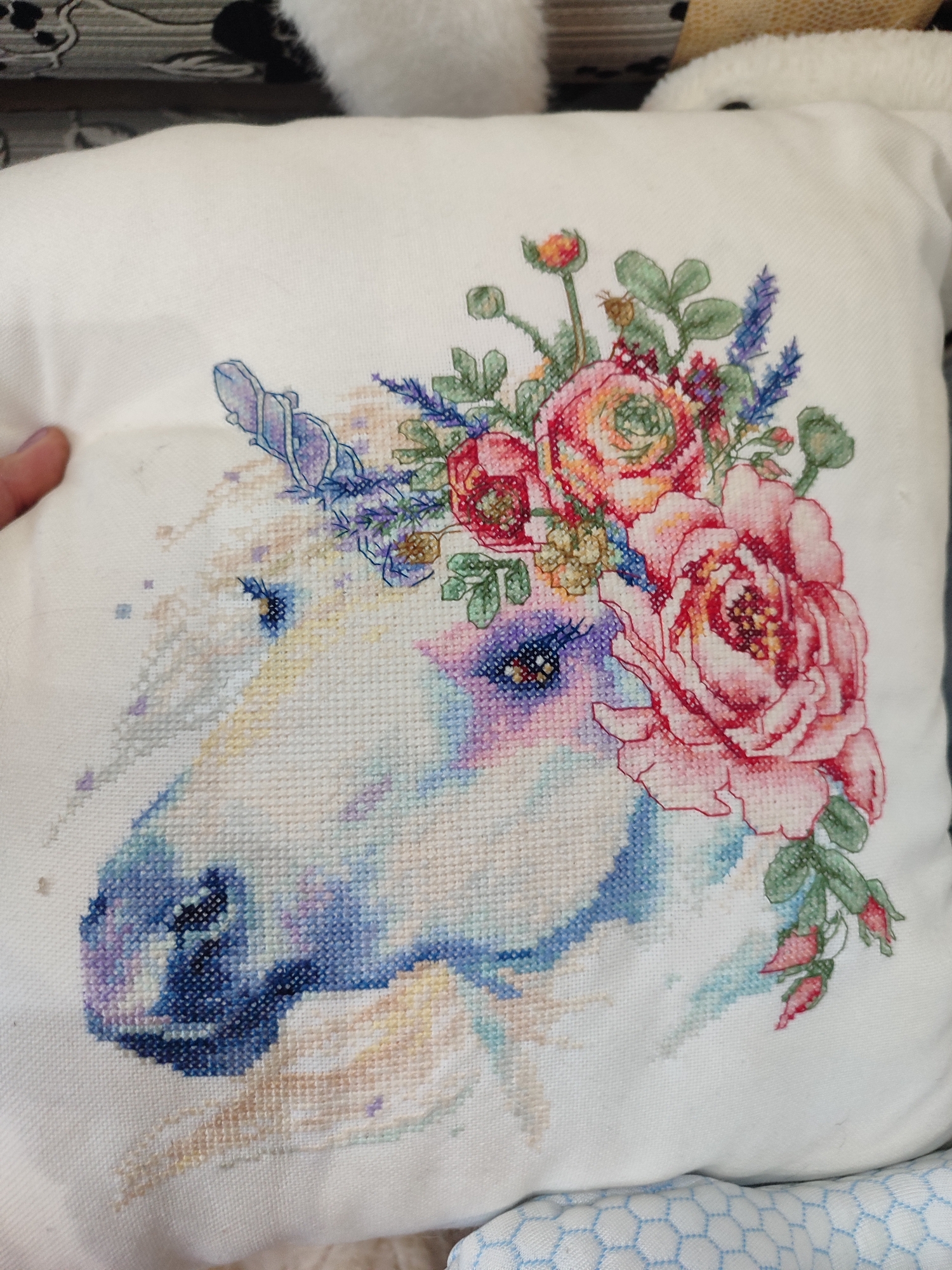 This media file showcases a close-up of a white pillow adorned with a detailed cross-stitch embroidery of a unicorn's head. The unicorn is depicted with a soft, ethereal quality, its mane transitioning into a vibrant floral crown. The flowers in the crown are rendered in rich shades of pink, red, and orange, with delicate green leaves and buds. Sprigs of lavender add a touch of blue to the floral arrangement.
The unicorn's face is a blend of soft blues, purples, and whites, suggesting a magical and gentle nature. Its eye is a striking deep blue, outlined with purple stitches. The embroidery is intricate, with individual stitches clearly visible, indicating it is a handmade piece. A human finger is visible on the left side of the image, lightly touching the pillow, giving a sense of scale and implying someone is holding or presenting the artwork. The background is softly blurred, with hints of furniture and fabric, suggesting the pillow is placed in a domestic setting, possibly a living room or bedroom. The lighting appears to be indoor, with no specific indications of time of day or weather. There is no visible text in the image. The overall impression is one of artistry and delicate beauty, highlighting the craftsmanship of the cross-stitch.