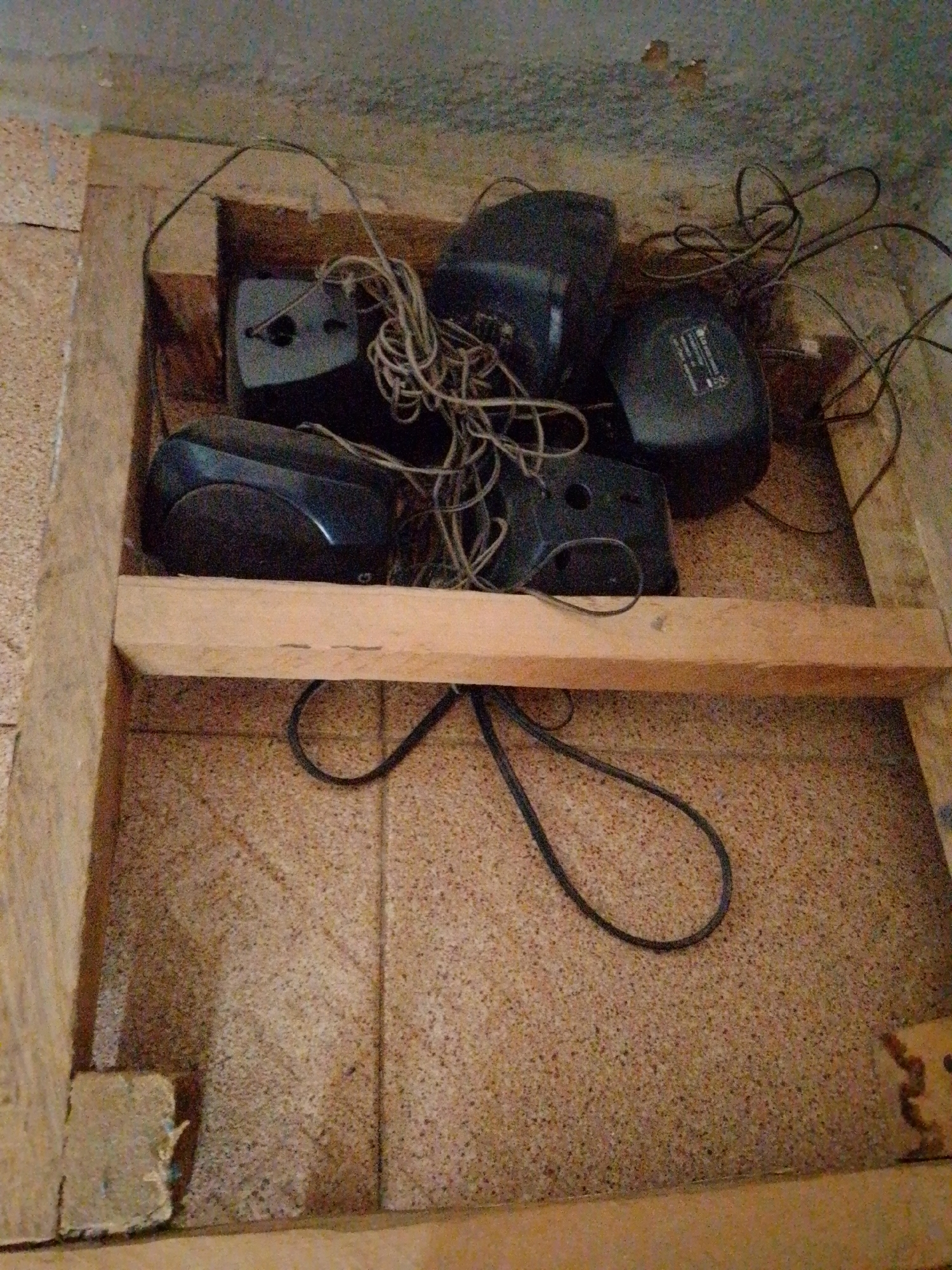 In a dimly lit, possibly unfinished or storage space with textured walls and tiled floor, four black electronic devices, resembling computer speakers or power adapters, are clustered together. They are connected by a tangle of wires. The devices are arranged within a crude wooden frame or enclosure. One device has a label with visible text. The overall scene suggests a utilitarian or storage area, possibly a basement or attic.
