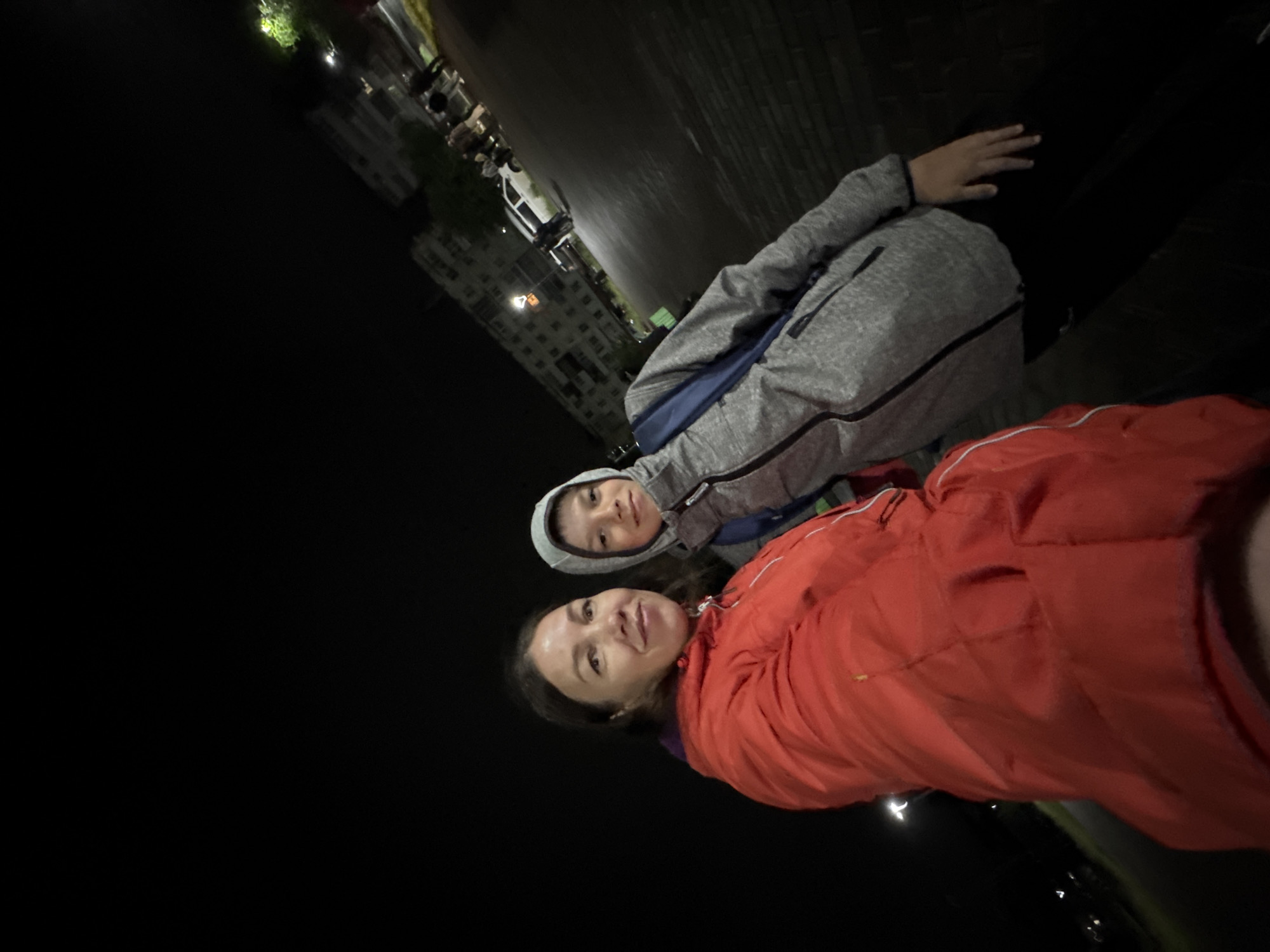 The image captures an adult female and a juvenile male posing for a selfie at night on a street in the city of Chaikovsky, Russia. The adult female, positioned on the left, has dark hair and is wearing a bright orange-red hooded jacket, revealing a purple lining near the neckline. She is looking directly at the camera with a slight smile. The juvenile male, positioned on the right, is wearing a grey hooded jacket with dark contrasting zippers and trim, and a blue backpack is visible over his right shoulder. He is also looking towards the camera with a slight smile. The background is illuminated by artificial light sources against a dark night sky. To the right of the subjects, a multi-story residential building with multiple lit windows is visible. In front of the building, a paved street or parking area contains several parked vehicles, including a white sedan, and indistinct human figures further in the distance. The immediate lower right foreground shows a dark, brick-patterned surface, consistent with a sidewalk or pedestrian area.