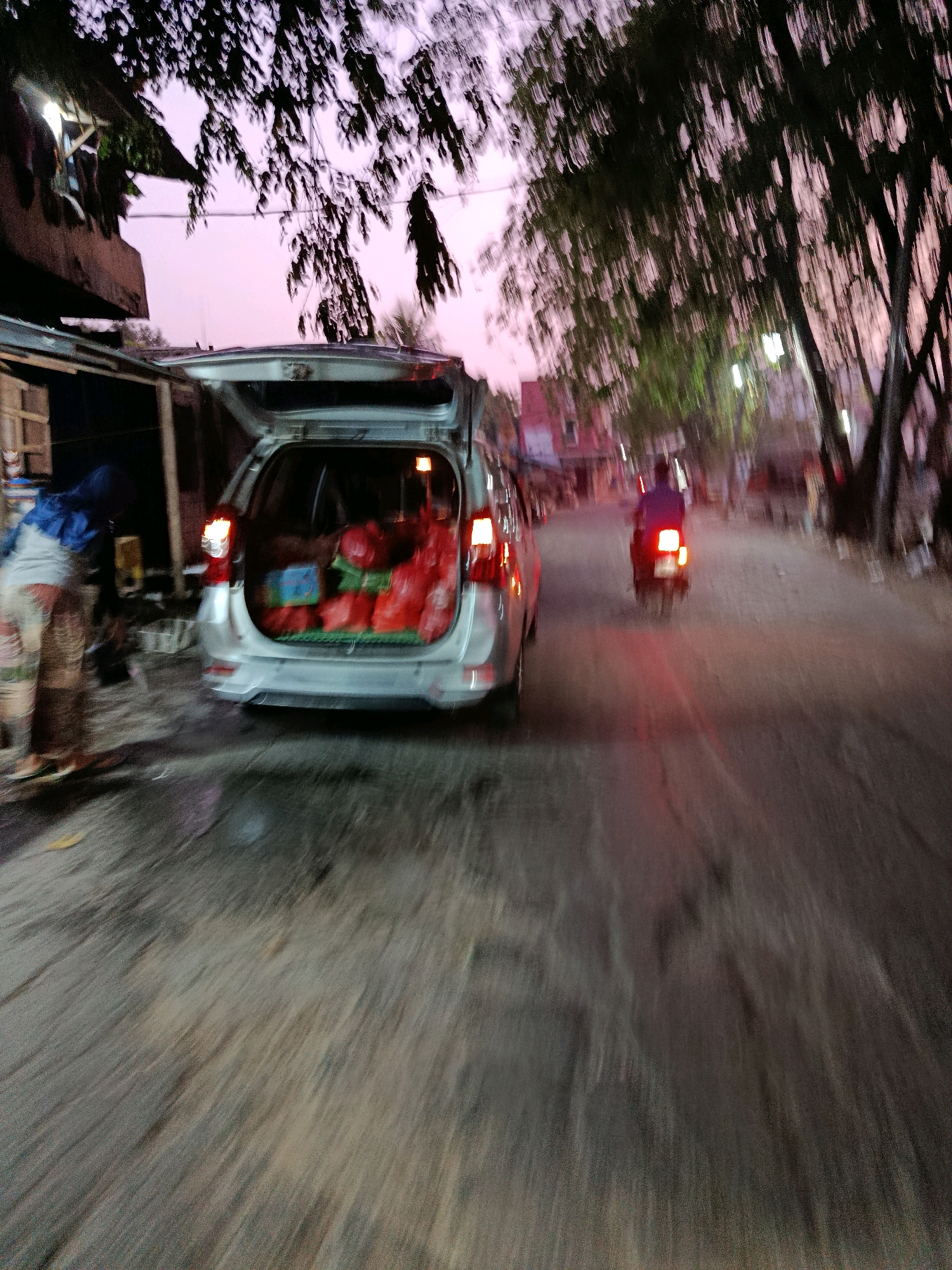 The image captures a street scene at dusk in Bekasi, Indonesia. A silver minivan with its trunk open is parked on the side of the road, laden with red bags and some blue boxes. To the left of the minivan, a person wearing a blue headscarf and patterned clothing is partially visible, appearing to be interacting with the contents of the vehicle. The street is unpaved and appears wet, with tire tracks suggesting recent traffic. A motorbike with a rider, illuminated by its red taillight, is moving away from the camera down the street. Trees with dense foliage overhang the scene, and streetlights are visible in the background, casting a soft glow on the surroundings. The sky is a gradient of soft pink and purple, indicative of twilight. The overall impression is one of a busy street at the end of the day, possibly a marketplace or residential area.