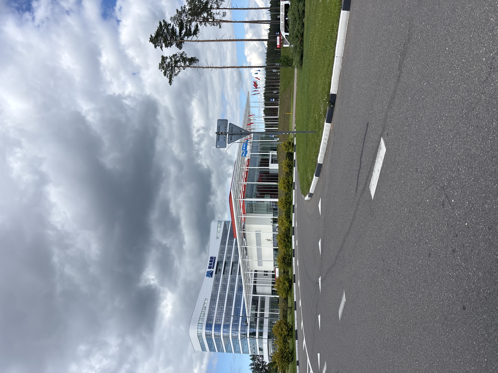 This image captures an exterior view of a modern, multi-story building with a curved facade and numerous windows. The building features a white exterior with blue and red accents. A prominent logo with blue text and a stylized blue triangle is visible on the upper left portion of the building, along with the text "BIOTECH" in blue. The building is situated next to a paved road with white markings and a black and white striped curb. Adjacent to the building is a well-maintained grassy area with decorative bushes. In the background, several tall trees stand against a sky filled with dramatic, heavy clouds, suggesting overcast weather. A line of flags on poles is also visible in the distance. The scene appears to be set in an open area, possibly a business park or industrial zone, given the architecture and the road infrastructure. The overall impression is one of a contemporary commercial or research facility under a moody sky. The text "BIOTECH" is clearly legible. The location is identified as Драчкаўскі сельскі Савет, Belarus. No people or specific activities are depicted.