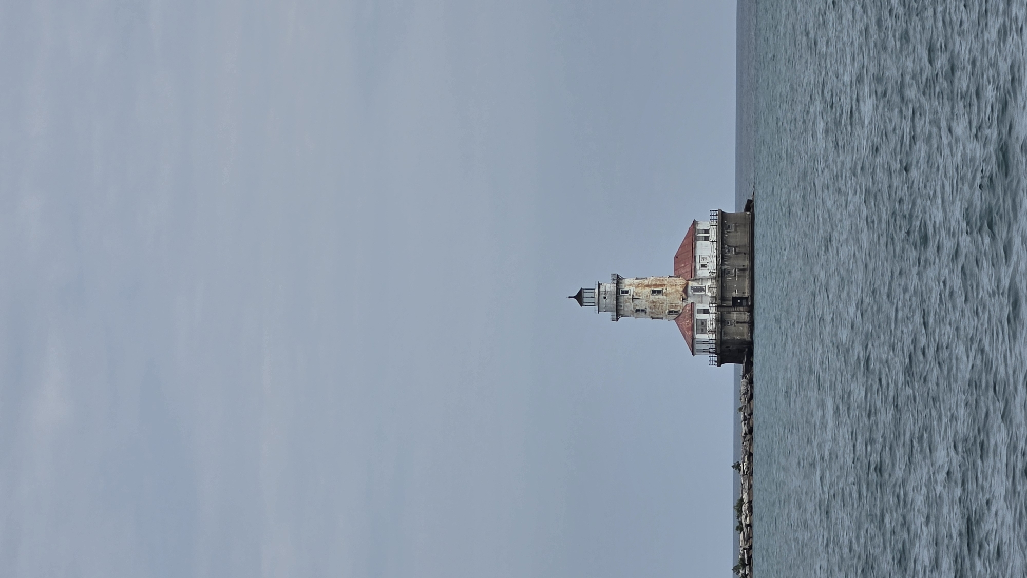 This is a photograph of the Chicago Harbor Lighthouse, situated in the waters of Lake Michigan off the coast of Chicago, Illinois, United States. The lighthouse, a white cylindrical structure with a conical top and a red roof, is perched on a stone foundation. It is surrounded by calm, dark blue water. A breakwater made of large stones extends from the land to the lighthouse, with a few small, sparse trees on the breakwater. The sky above is overcast and a pale blue-gray. There are no people visible in the image. The overall scene is one of a solitary lighthouse in a vast body of water under a muted sky.