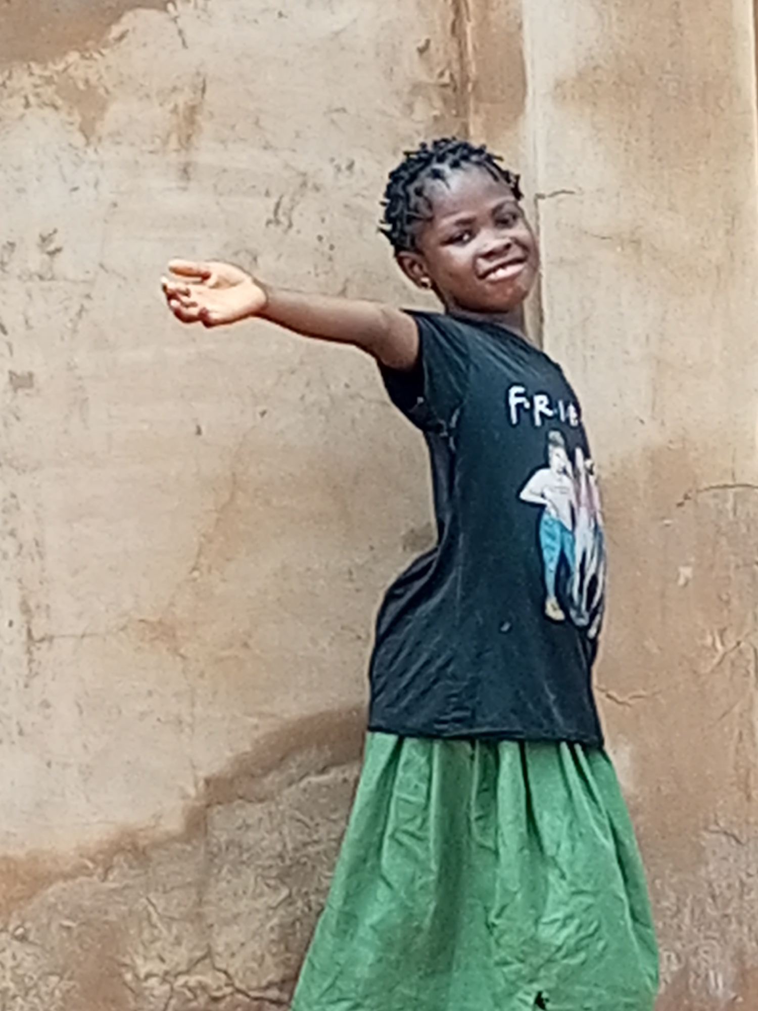 A young girl with braided hair and a black t-shirt with an image of two people and the word "FRIENDS" printed on it, and a green skirt, smiles at the camera. She stands in front of a textured, light brown wall. Her right arm is extended, palm open. The background appears to be an outdoor urban environment in Tempure, Nigeria.
