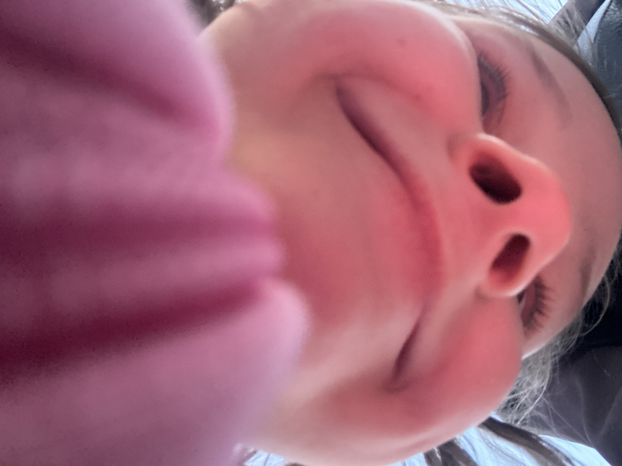 A close-up, low-angle photograph shows a person's face with a pink garment partially obscuring the left side of the frame. The visible features of the face include a nose, eyes, and mouth. The person appears to be smiling. The environment and location are undeterminable.
