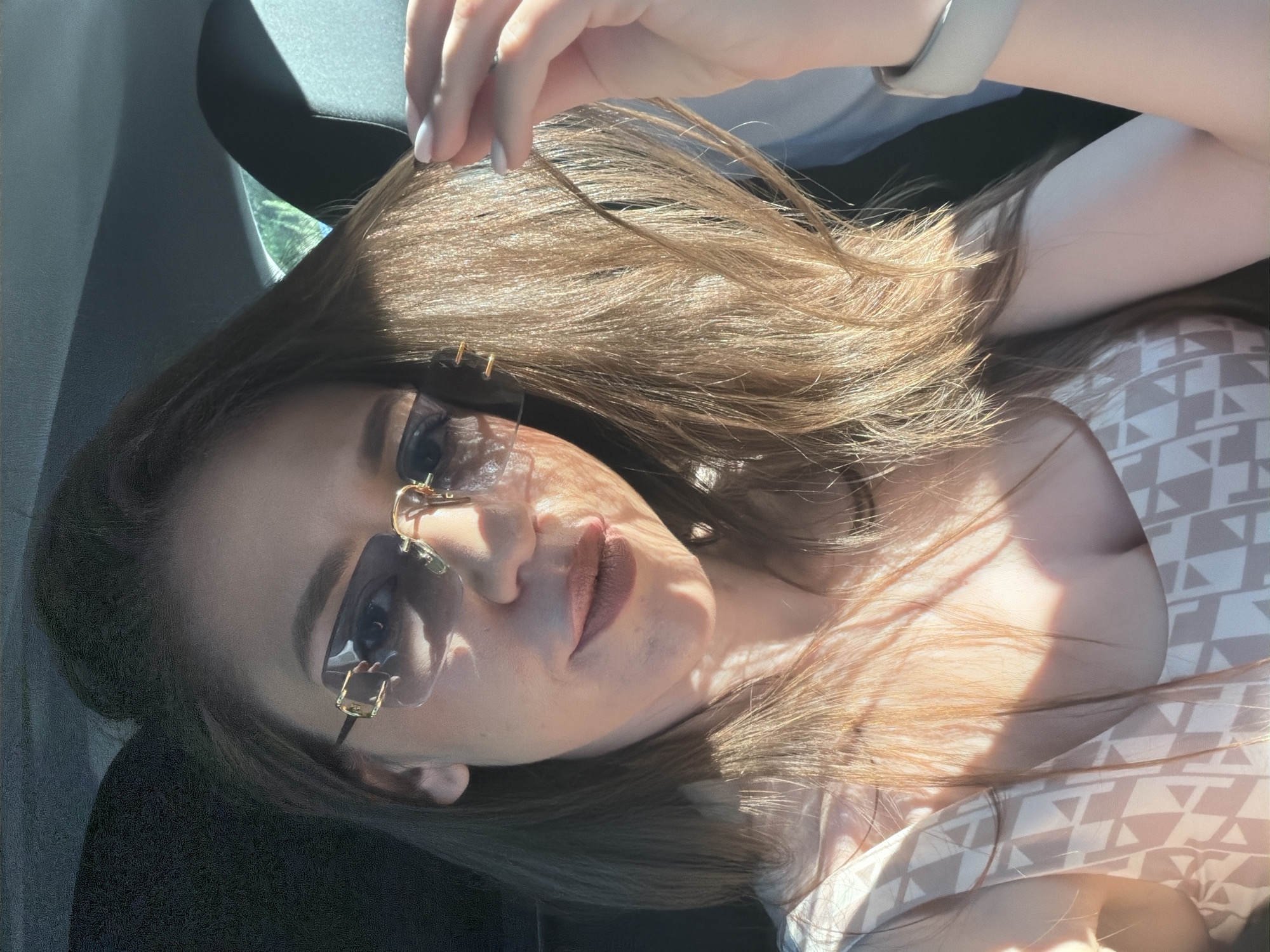 In the city of Sofia, Bulgaria, a woman with long, light brown hair is seated in a vehicle, likely a car, as indicated by the dark interior and a visible headrest. She is wearing stylish, rimless sunglasses with gold detailing on the frames and a patterned top. Sunlight streams in, casting shadows across her face and hair, highlighting a subtle makeup application on her lips. Her right hand is raised, gently touching her hair. A light-colored smartwatch is visible on her left wrist. The scene appears to be a candid shot, possibly taken while traveling.