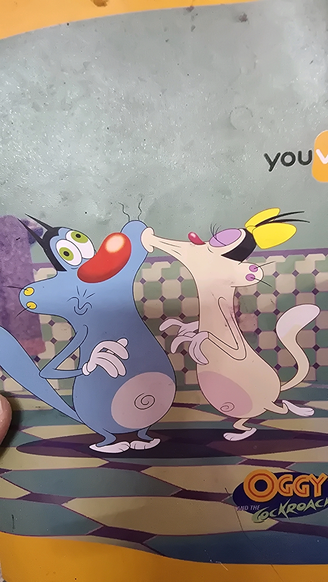 The image depicts a cartoon illustration featuring two anthropomorphic cats, presumably characters from "Oggy and the Cockroaches." The blue cat on the left has its eyes wide open and its nose touching the pink cat's mouth. The pink cat on the right has its eyes closed, its nose pushed against the blue cat's, and its tongue extended. Both cats have their arms outstretched, with their hands in claw-like positions. They are positioned in front of a background with a green and white checkered pattern, and a floor with a geometric design of green, yellow, and purple shapes. At the bottom right corner, there is a logo that reads "OGGY AND THE COCKROACH". Above the cats, partially visible on the right side, is the word "you" followed by an orange shape. The image appears to be a print on a textured surface, possibly a notebook cover or a placemat, with some wear and tear visible, including scratches and smudges. The overall impression is that of a still from a cartoon. There is no indication of this image being located in Bengaluru, India; the description is purely based on the visual content.