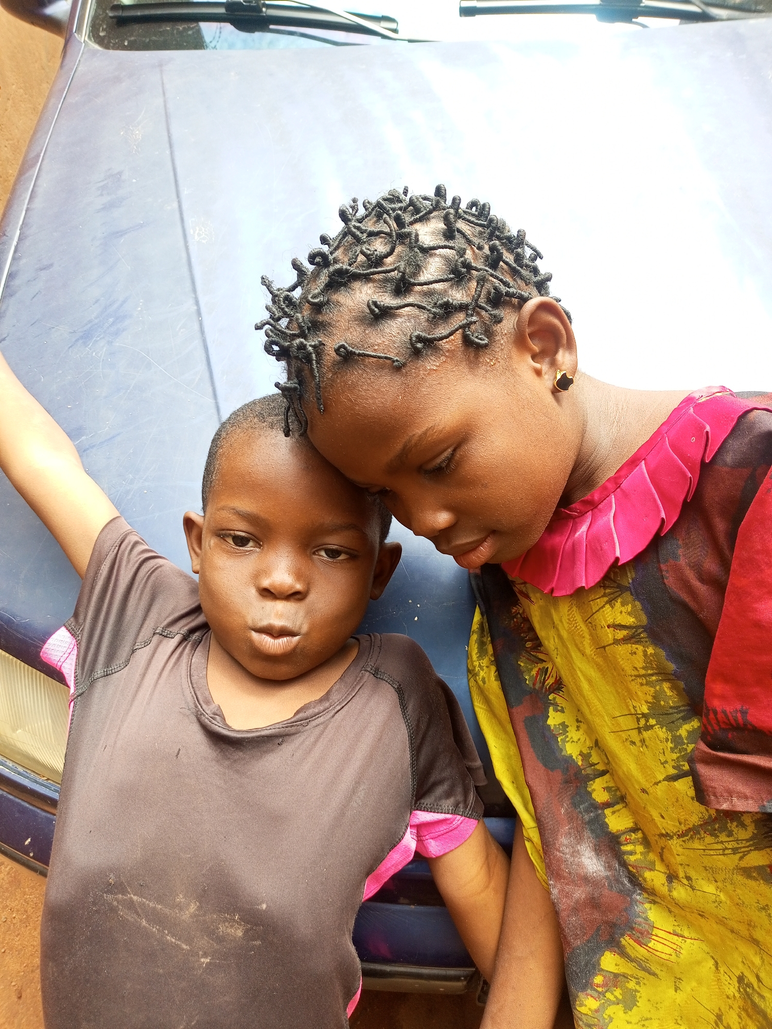Two young children, one older and one younger, are positioned close together, leaning against the hood of a dark blue vehicle. The older child, on the right, has dark, intricately braided hair and wears a colorful shirt with yellow, red, and black abstract patterns and a ruffled pink collar. This child is looking downwards, with their head tilted. The younger child, on the left, is wearing a dark brown t-shirt with pink trim. This child is looking directly at the camera with a slightly pursed expression and their left arm is raised. The background consists of the vehicle's hood, which appears dusty, and the suggestion of an outdoor environment. The location is identified as Tempure, Nigeria.