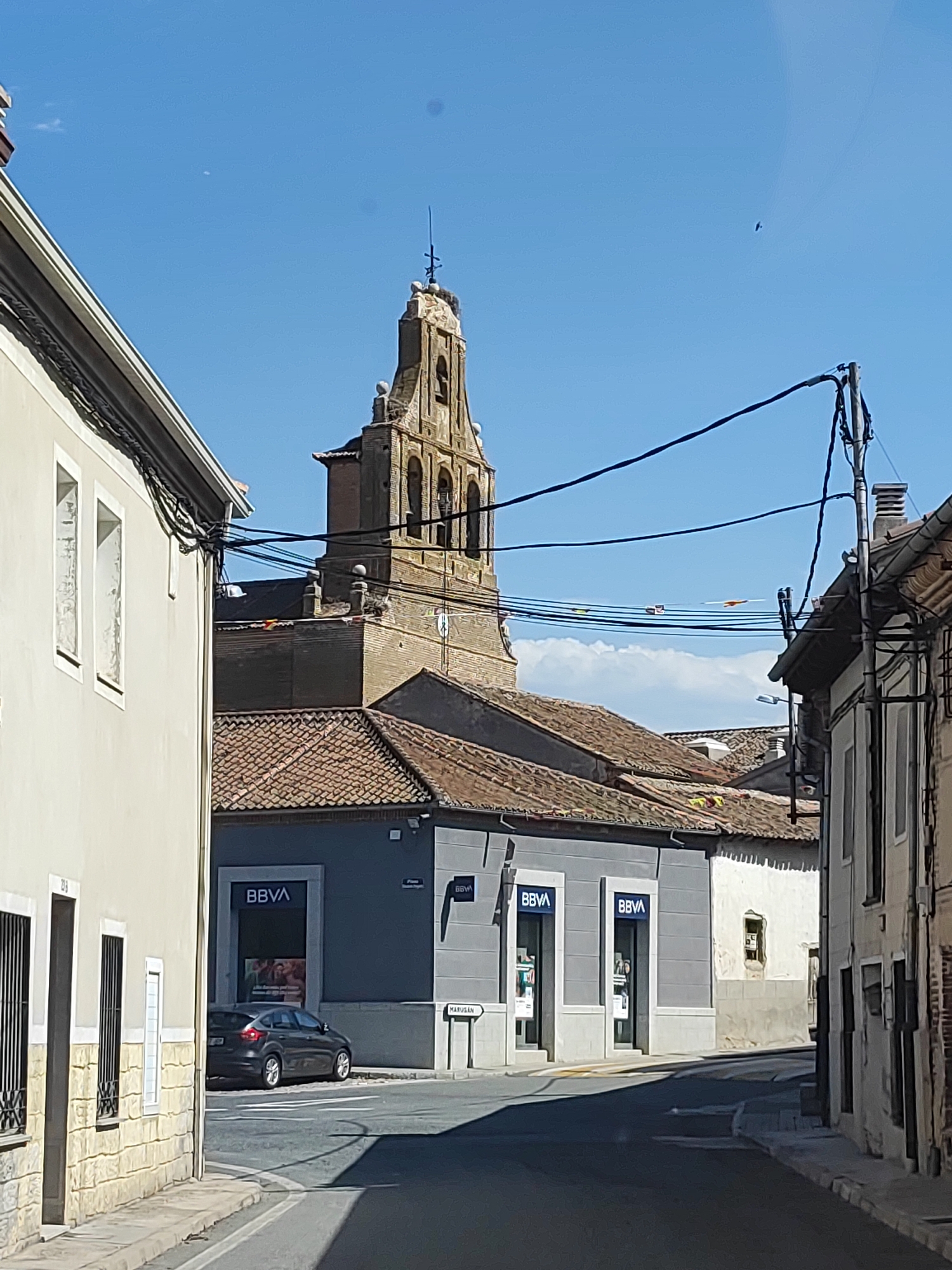 The image captures a street scene in Sangarcía, Spain, dominated by traditional Spanish architecture and a clear blue sky. In the foreground, a gray car is parked on a paved street that curves to the right. The car is facing away from the viewer and towards a building with a dark gray facade featuring the blue and white "BBVA" logo twice, indicating a bank. Adjacent to this building, another structure with a lighter, textured facade is visible.
Further into the background, a prominent church tower with arched openings and a cross on top stands against the sky. The tower is constructed of weathered brick and stone. The roofs of the buildings in the middle ground and background are covered in terracotta tiles. Electrical wires crisscross the sky, with some attached to poles and others running between buildings. A street sign with the word "MARUGAN" is visible on a white post to the right of the frame. The overall impression is of a quiet, sunny day in a historic Spanish town.