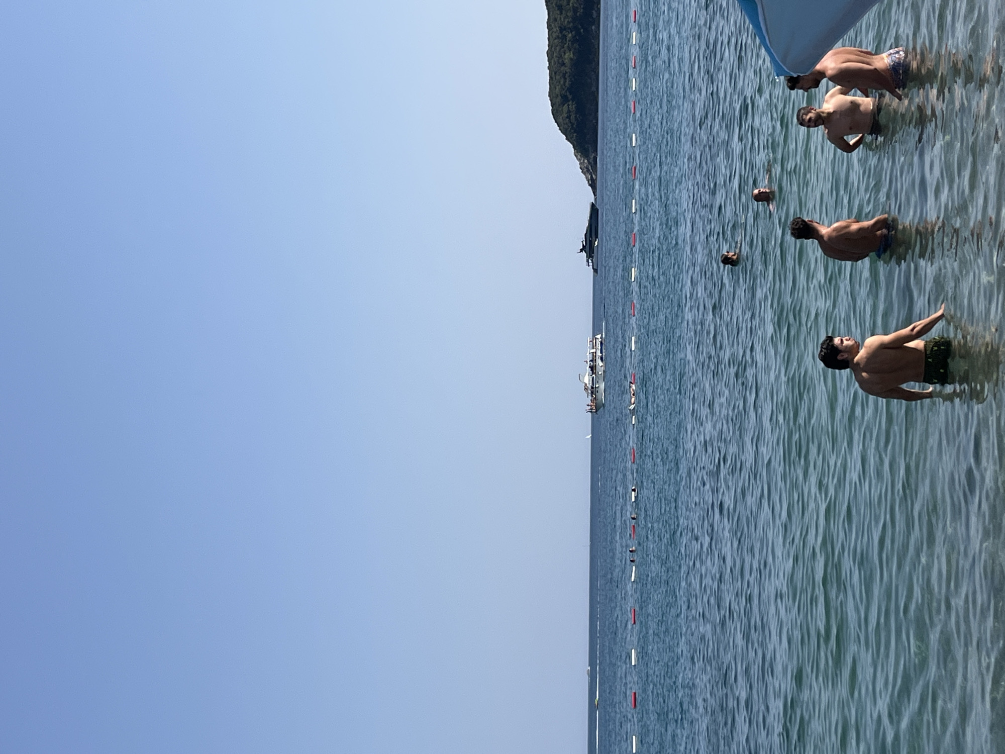 The image depicts a coastal scene at a beach in Prijevor, Montenegro, on a clear, sunny day. The calm, blue water of the Adriatic Sea fills the foreground and mid-ground. A line of buoys, alternating red and white, marks a designated swimming area. In the distance, several boats are visible on the water, including a white passenger boat and a larger, dark yacht. A treed headland juts out into the sea on the right side of the image.
Several individuals are present in the water. In the foreground, four men are standing in the shallow water, some interacting with each other. Further out in the water, other swimmers can be seen. One person appears to be relaxing on an inflatable in the distance, near the buoy line. The overall atmosphere is one of leisure and enjoyment of a summer day at the beach.