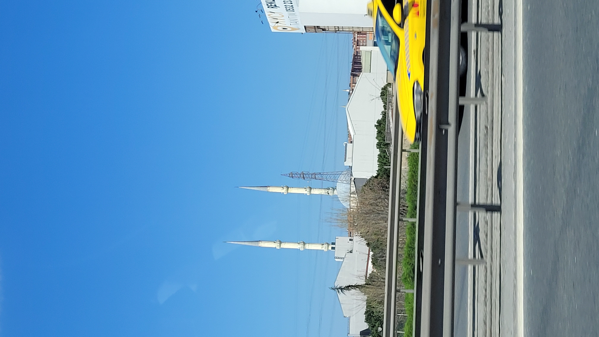 The image captures a roadside view in Küçükçekmece, Turkey. Two prominent minarets of a mosque stand against a clear blue sky. An electrical transmission tower is situated between them. To the right of the minarets, there is a building or complex of white structures. A portion of a yellow vehicle is visible at the top right corner of the frame, partially obscured by a roadside barrier consisting of grey metal posts and rails. The foreground is dominated by an asphalt road surface with painted lines.