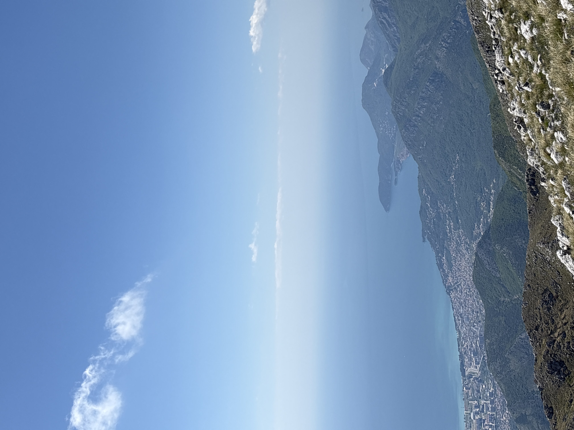The image shows a view from a high vantage point in Mali Mikulići, Montenegro. The scene includes a body of water, likely a lake or part of the Adriatic Sea, a town or city built along the coast, and the rugged terrain of the mountains. The foreground includes rocky, grass-covered terrain. The sky is a clear blue with a few scattered clouds. The overall impression is that of a scenic landscape viewed from a high altitude.