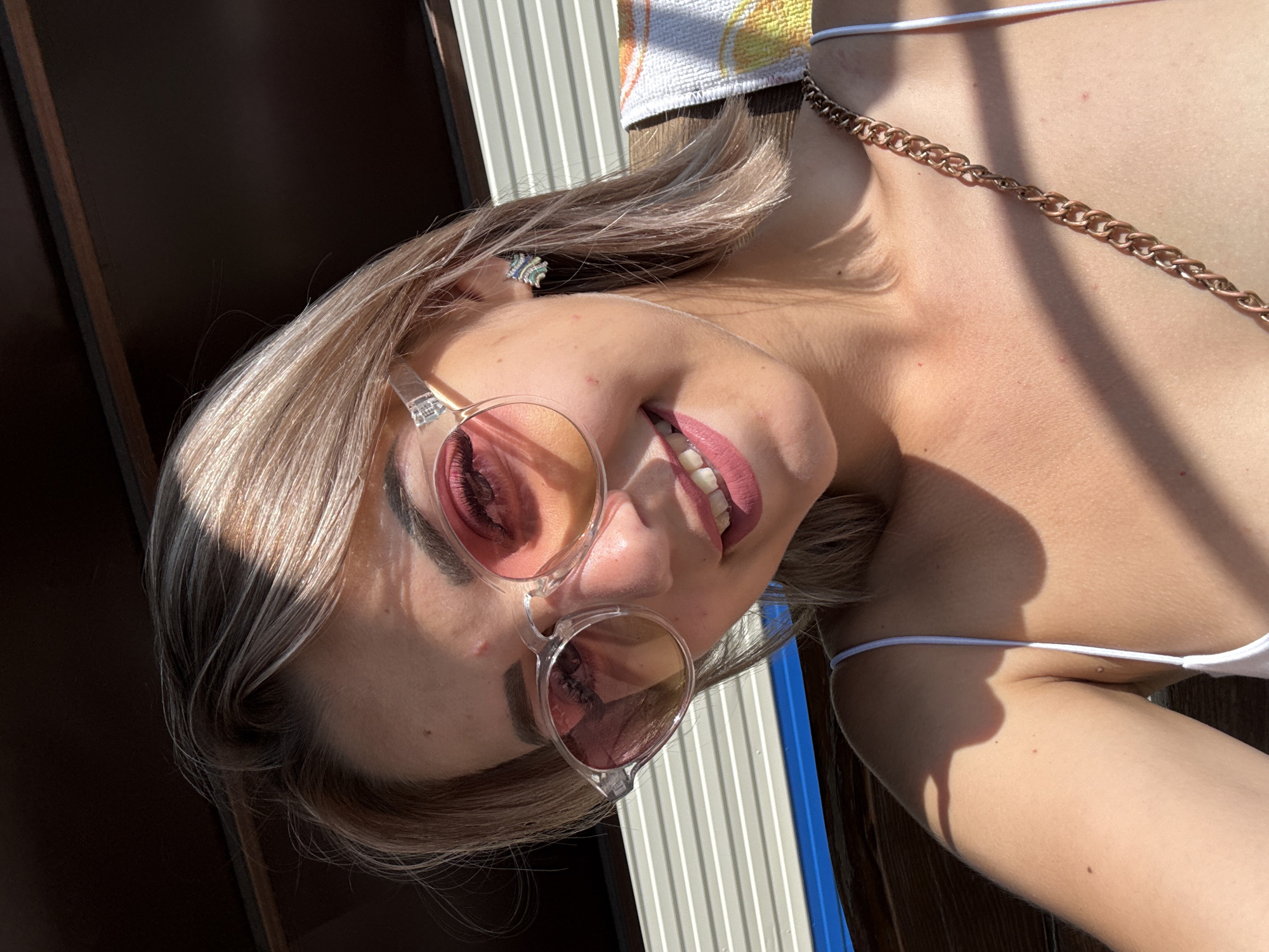 A young woman with blonde hair and pink-tinted sunglasses is outdoors, likely in Krasnogorskoye, Russia. She is wearing a white tank top and a gold chain necklace. Her expression is a slight smile, and she is looking directly at the camera. The background appears to be a slatted structure, possibly a porch or a cabana, with a towel draped over it. Sunlight casts shadows across her face and chest.