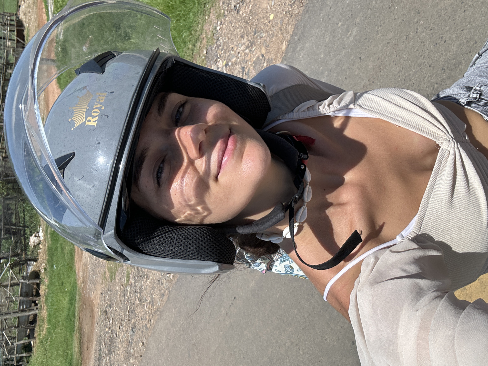 A woman wearing a grey helmet with a gold "Royal" emblem and a clear visor is looking directly at the camera. She is wearing a light-colored, sheer long-sleeved top over a ribbed white tank top and denim shorts. Around her neck, she has a necklace made of white shells. She is outdoors on a paved road, with a grassy area and some wooden structures in the background. The scene appears to be in a rural or semi-rural area.