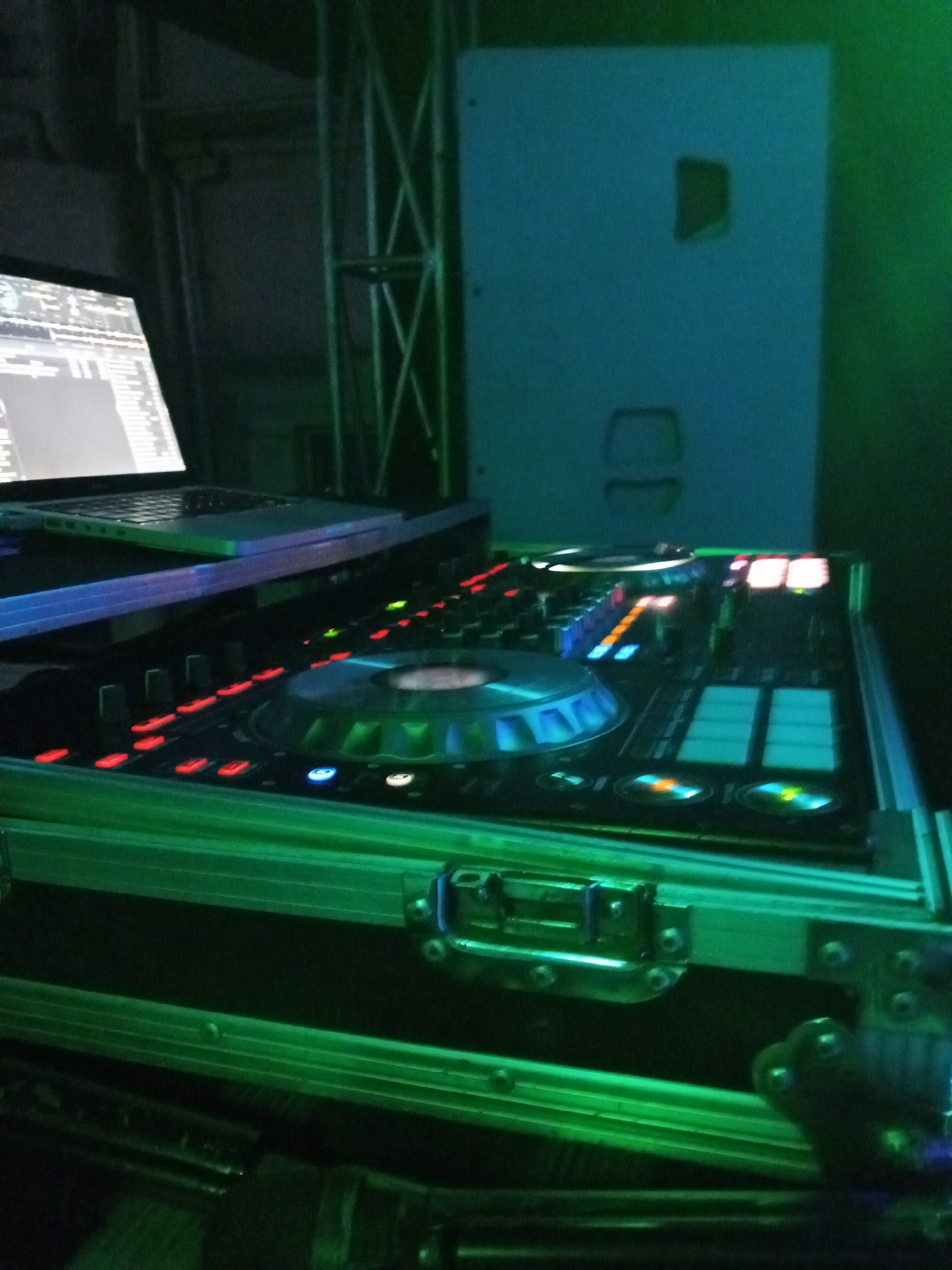 The image depicts DJ equipment, including a laptop and a DJ controller, set up in a dimly lit environment. The equipment is housed in a flight case with metal hardware. The background includes a speaker and some structural support. The lighting is predominantly green. There are no people visible. The location is unknown, so it cannot be confirmed if the image was taken in Lokoja, Nigeria.