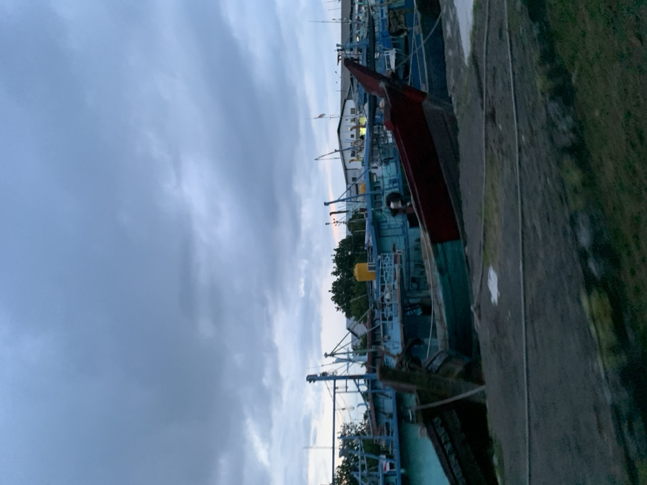 The image shows a scene at a harbor in Probolinggo, Indonesia, a city located at latitude: -7.731, longitude: 113.218. The image shows several boats, with a prominent red and blue boat in the foreground, and a smaller boat in the background. All the boats are docked in the harbor, and there are no visible people present. The sky is overcast with a dark, grey color, and the water is calm. The boats are tied together and are facing away from the viewer. There is a green grassy patch of land in the bottom right corner of the image.