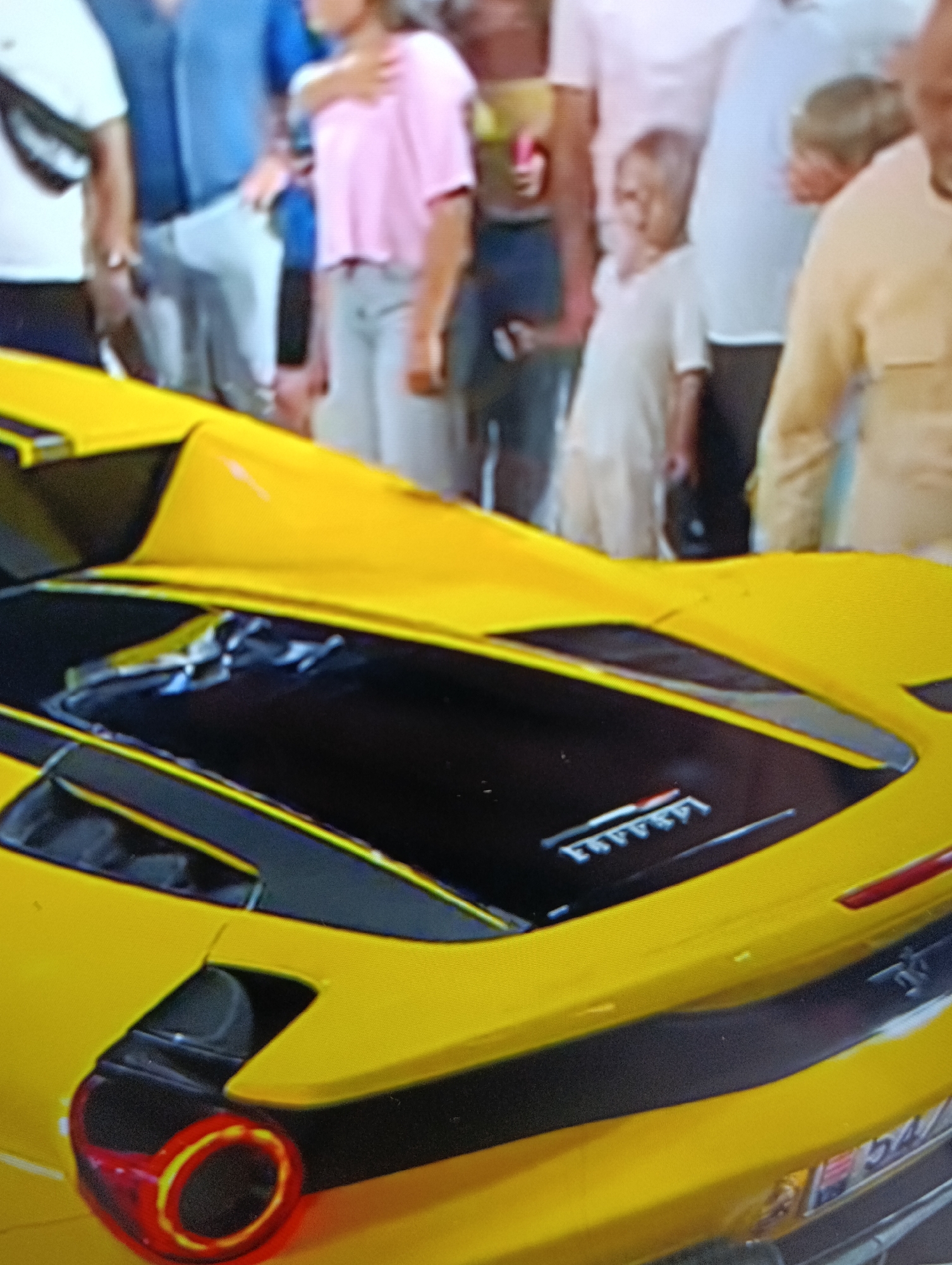 The image shows the rear of a yellow Ferrari car, with the "Ferrari" logo visible. The car has a black engine cover and round taillights. Behind the car is a crowd of people of various ages and genders. The scene appears to be outdoors in a city environment, though the specific location is unknown.