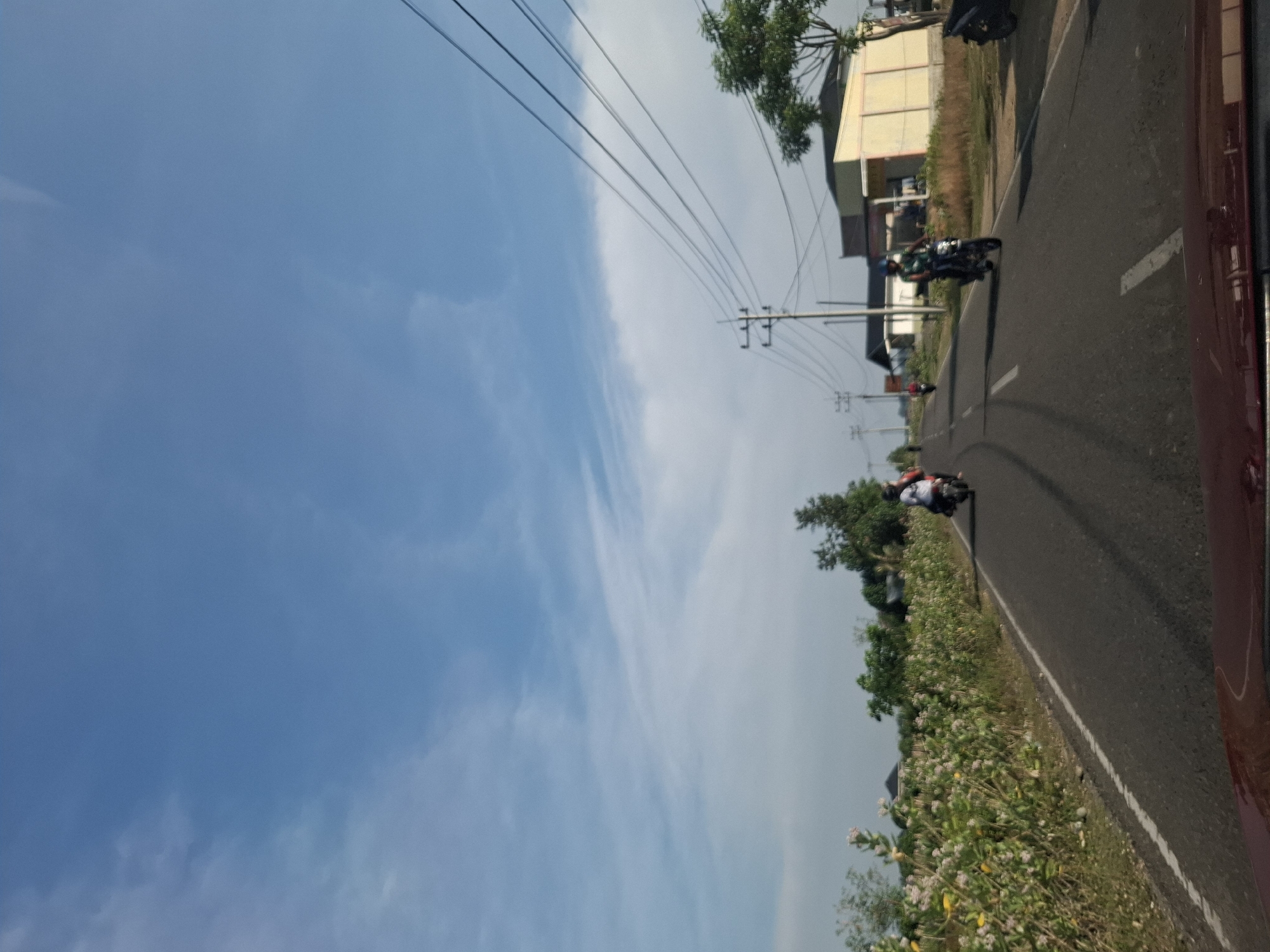 The image depicts a rural road scene in Sukon, Indonesia, under a bright blue sky with scattered white clouds. The road, paved with asphalt and marked with white lines, curves gently into the distance. On the left side of the road, a dense growth of flowering bushes with white and yellow blooms lines the roadside. On the right, a grassy area borders the road, leading to a building with a light yellow facade and a dark roof, accompanied by utility poles with multiple power lines strung across. Several individuals are seen riding motorcycles on the road. One motorcycle, with two riders, is in the middle distance on the left side of the road. Further ahead, a single rider on a motorcycle is visible. The image appears to be taken from a vehicle, as indicated by a portion of a dark red car interior at the bottom of the frame.