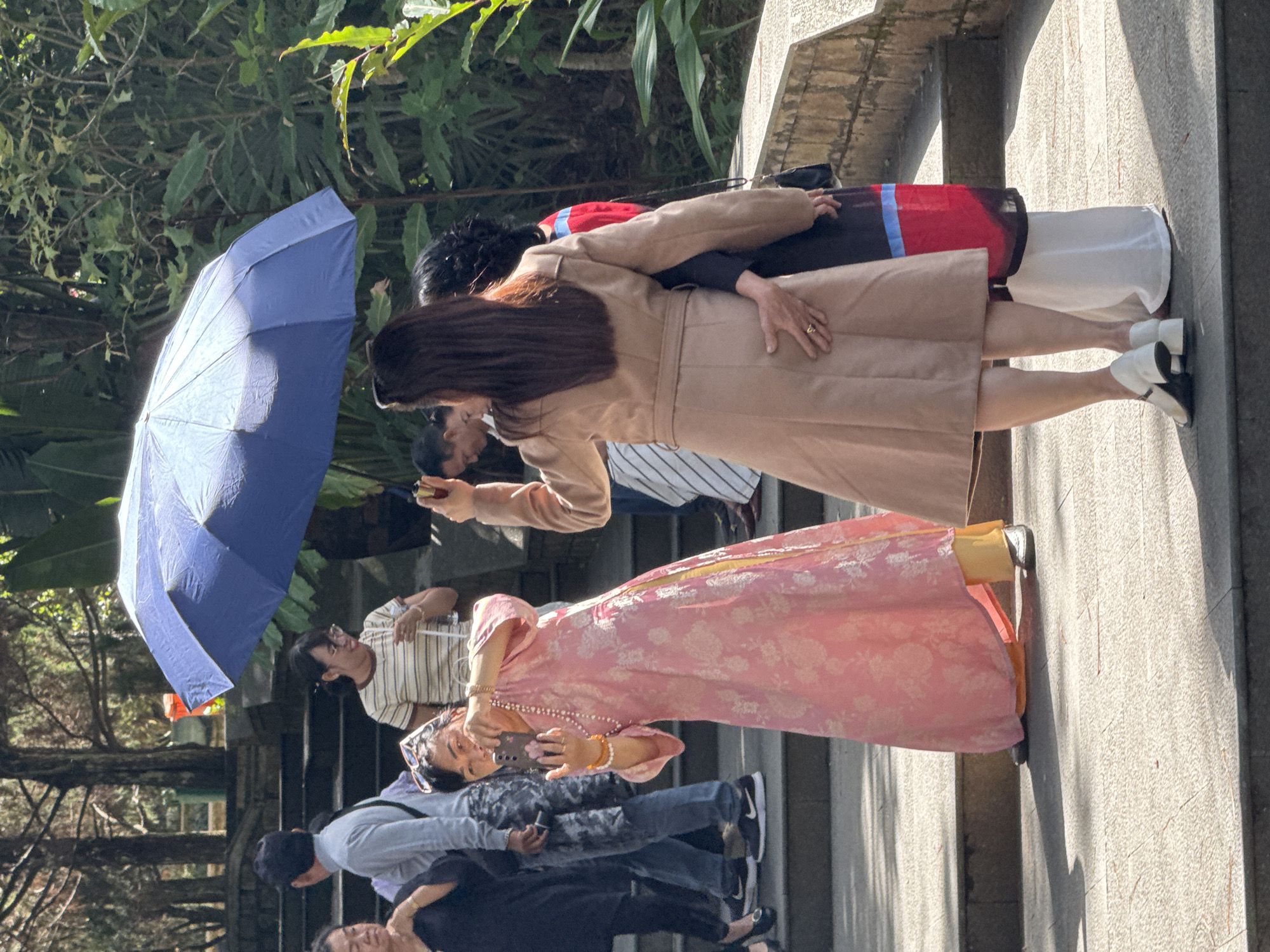 This is a photograph of a group of people on a set of stairs, likely in a park or tourist attraction in Da Lat, Vietnam. The central focus is a woman in a long, pink, floral patterned dress and yellow pants, holding up a smartphone to take a selfie. She is adorned with bracelets and a pearl necklace. To her right, another woman in a tan coat is standing, with her arm around the woman in the pink dress and her hand resting on the latter's lower back. This woman is also wearing a ring. Behind these two women, another woman is visible, wearing a black garment with red and blue stripes. A blue umbrella is held open above the group, providing shade. In the background, several other individuals are present, some walking up the stairs, and a man in a light grey shirt and dark pants is clearly visible. The environment appears to be outdoors, with lush green foliage and trees visible in the background, suggesting a natural setting within the city. The stairs are made of stone or concrete, and the ground is paved with tiles. The lighting indicates it is daytime, with strong sunlight casting shadows.