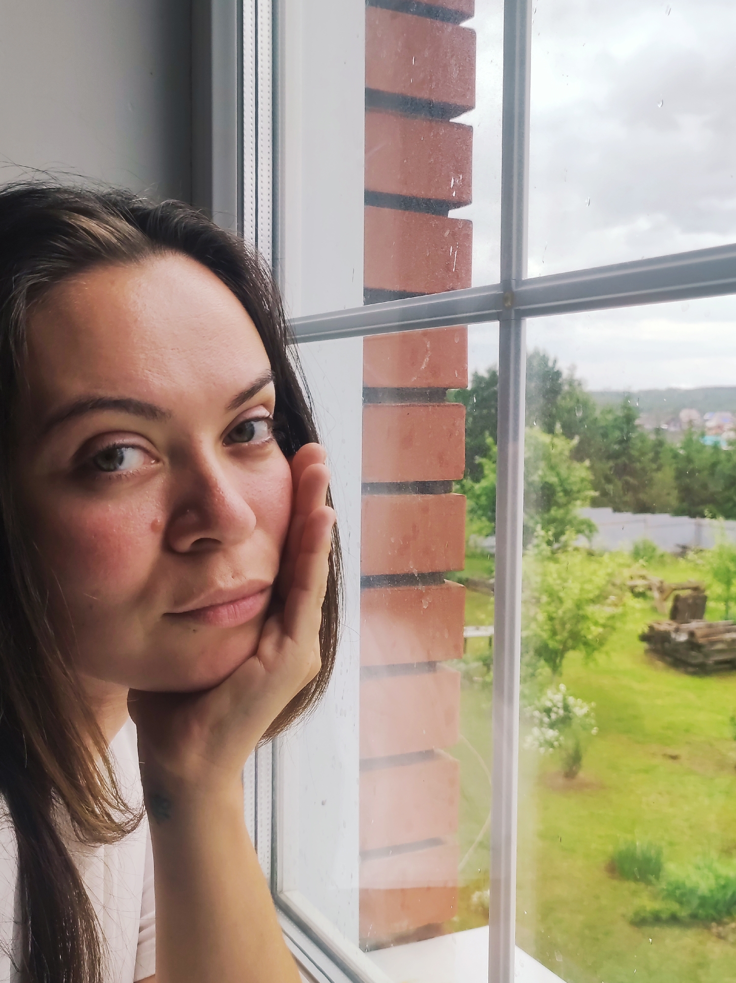 A close-up, eye-level indoor shot shows a woman looking out a window, her face resting on her right hand. She has dark hair, a light complexion, and dark eyes that gaze slightly to the right. Her expression appears calm and contemplative. She wears a light-colored top. A small, dark, possibly abstract tattoo is visible on her left wrist, just above her hand.
The window features a white frame and mullions, dividing the view into panes. Raindrops are visible on the window glass, suggesting it is raining or has recently rained. Through the window, an outdoor scene unfolds. To the left, directly outside the window, a reddish-brown brick wall is visible, indicating the exterior of the building. Beyond the immediate foreground, there is a vibrant green landscape with numerous trees and grass. A white fence can be seen in the midground, and in the distance, what appears to be scattered houses are visible against an overcast, grey sky. Some stacked wooden elements, possibly pallets or logs, are also present in the outdoor scene. No text is visible in the image.