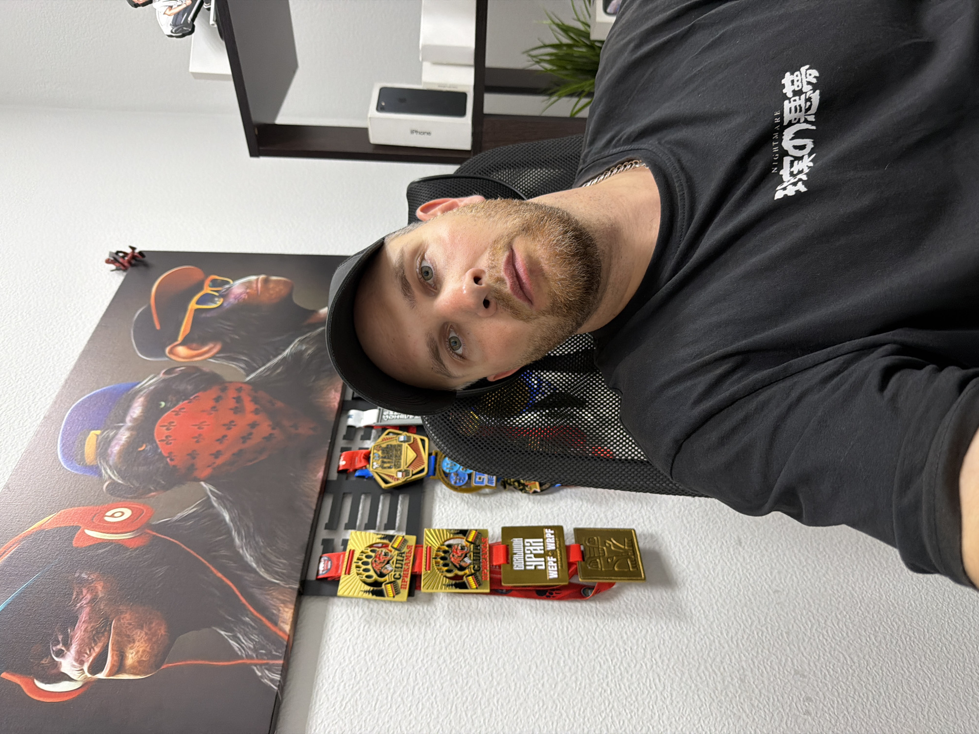 The image shows a man sitting in a black office chair in front of a wall. He is wearing a black t-shirt and a black baseball cap. Behind him, a large framed picture of three stylized monkeys is hung on the wall. To the left of the man, a rack with several medals is mounted on the wall. The medals are gold and red, with some inscription visible on them. To the right of the man, a dark brown shelving unit is visible, holding a black iPhone in its box and a small green plant. The wall has a textured surface. The man appears to be looking towards the camera with a neutral expression. There is no clear indication of the specific location within the city of Chaikovsky, Russia, however, the medals bear inscriptions in Cyrillic, which is the alphabet used in Russia.