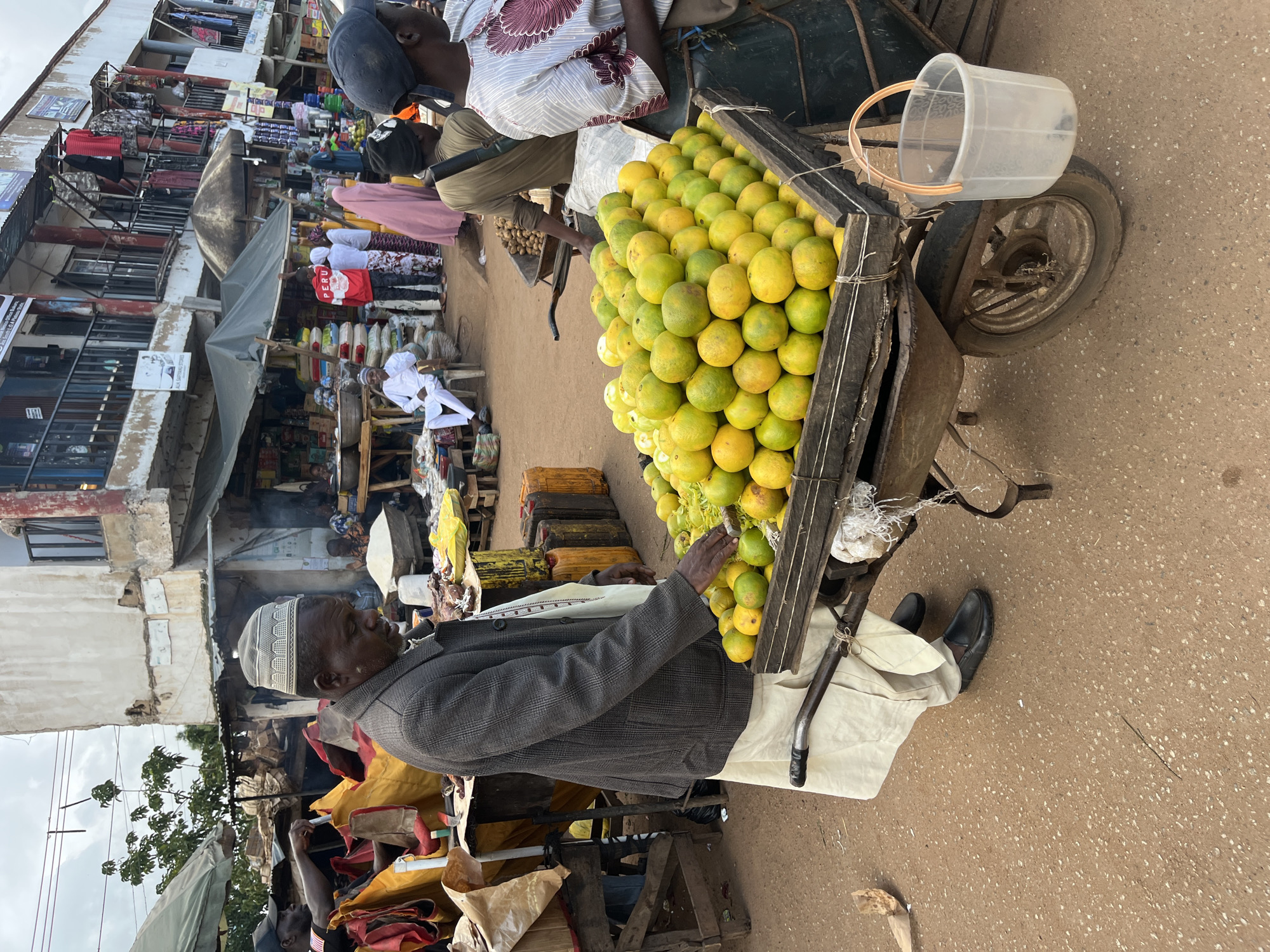 The image depicts a street market scene in Bukuru, Nigeria. The primary subject is a man wearing a grey plaid jacket and a white kufi, seated next to a cart laden with a large quantity of oranges. The cart is a wooden structure on wheels, with a metal frame and a clear plastic bucket on top. The man appears to be selling the oranges.
In the background, there are various market stalls with goods displayed, including clothing and packaged items. Other people are visible in the distance, some appearing to be vendors or shoppers. The setting is an outdoor market with a dirt or gravel ground. The overall atmosphere suggests a bustling marketplace.
Notable details include the abundance of oranges, the rickety nature of the cart, and the man's attire, which includes traditional Islamic headwear. The lighting suggests it is daytime, possibly late morning or afternoon, given the shadows and general brightness. There are no explicit cues regarding the weather, but the ground appears dry.
The image is oriented vertically, with the ground appearing on the right side of the frame and the market stalls on the left. The focus is primarily on the man and the oranges, with the surrounding market serving as context. There is some visible text on posters and signs in the background, but it is not legible.
