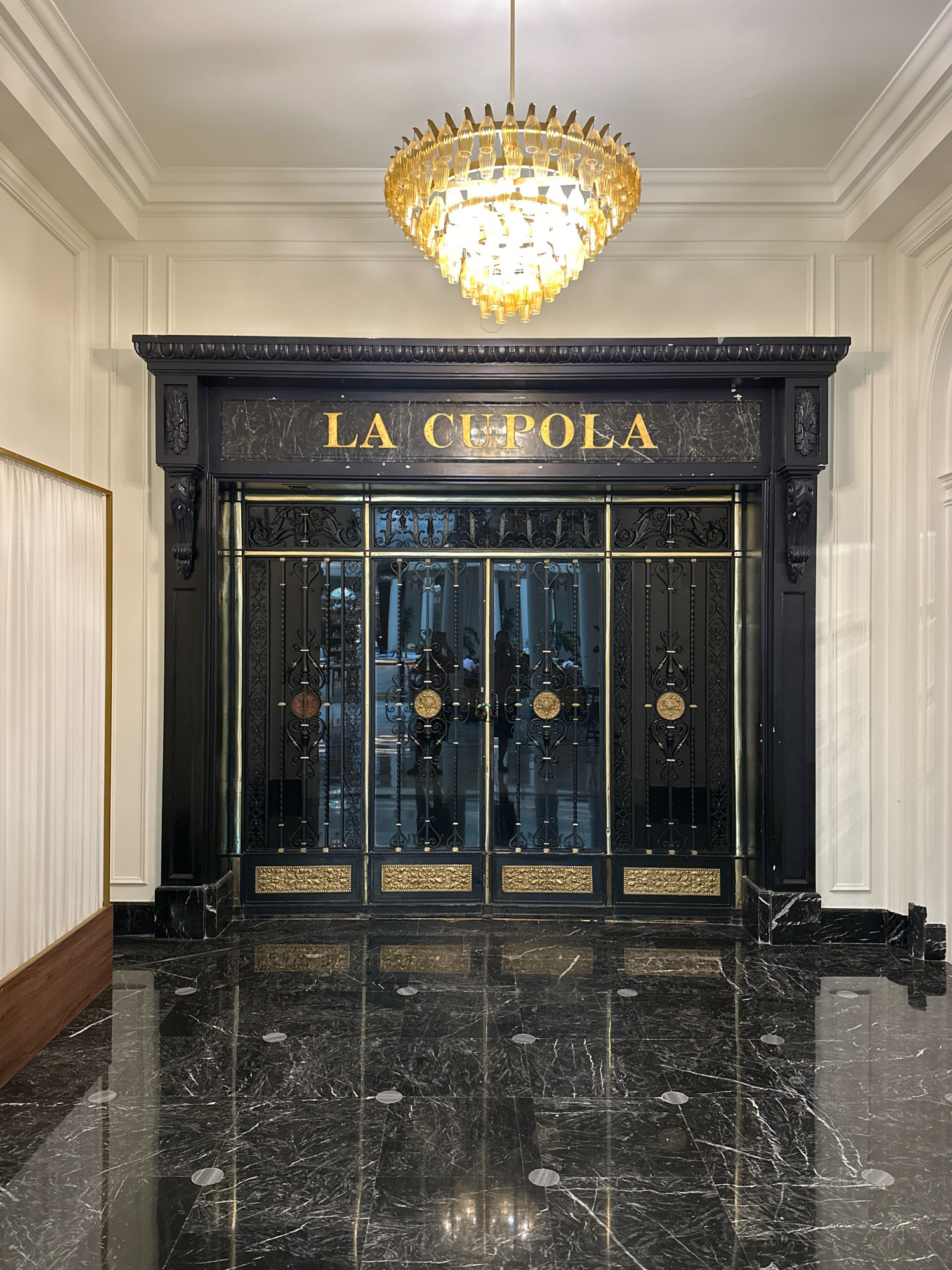 The image shows the entrance to "LA CUPOLA" restaurant, located in Madrid, Spain. The entrance features ornate black metal doors with gold accents and the restaurant's name emblazoned above in gold lettering. A large, modern chandelier with amber-colored glass hangs from the ceiling. The floor is made of polished black marble with white veining, and it reflects the chandelier and the entrance. To the left of the entrance, a white curtain with a golden frame is visible. The overall aesthetic is one of luxury and sophistication.