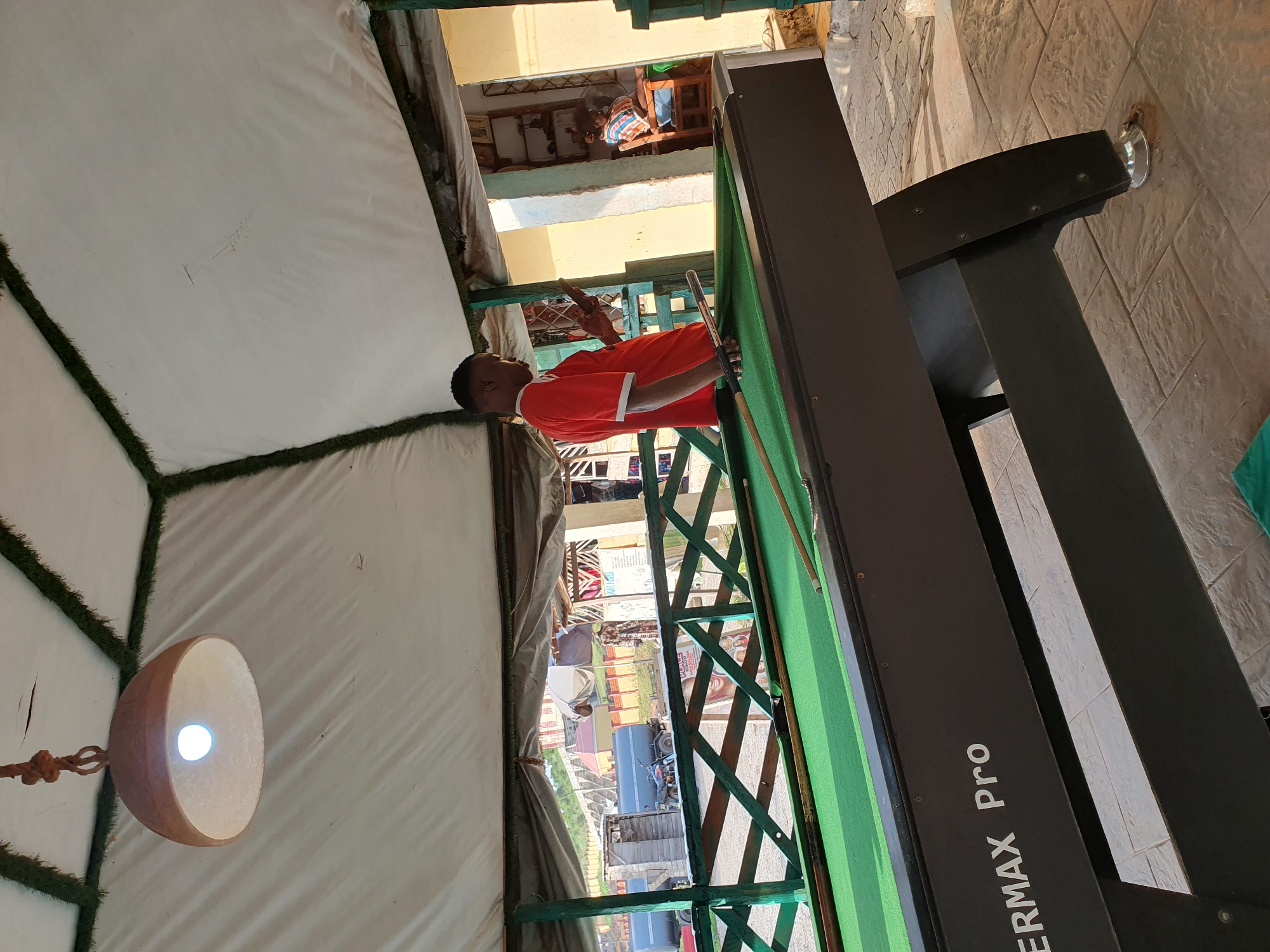 A young Black man in a red t-shirt and white trim stands by a pool table, looking at his phone. He is in an open-air covered area, possibly an outdoor seating or recreation space. The pool table is dark with a green felt surface, and cues are resting on it. In the background, a wooden railing with green trim separates the area from a street scene with vehicles and buildings. A child in a checkered shirt sits on a chair in a room visible through an opening. The ground is paved with textured tiles. The overall impression is of a casual, possibly commercial, establishment in a warm climate, likely Lokoja, Nigeria, given the context of the location.