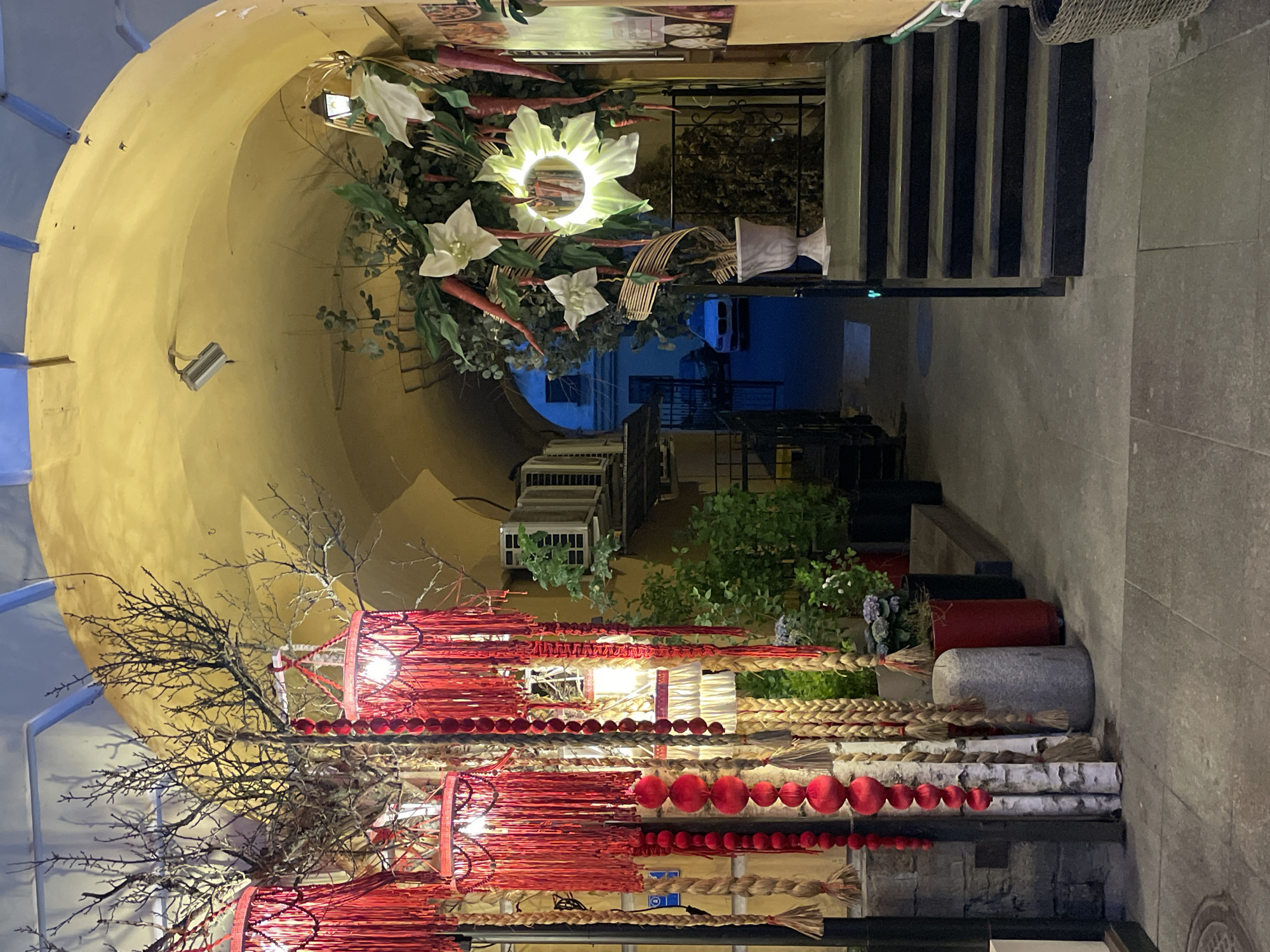 The image displays an ornately decorated arched passage, likely an entrance or alleyway, captured at night or during twilight. The interior of the archway is painted a warm, pale yellow.
In the foreground, a vibrant and eclectic array of decorations hangs and stands. This includes bare, branched tree limbs, intermingled with long strands of red woven material, some forming decorative balls. Braided natural fibers, possibly straw or rope, also hang vertically. At the base, segments of white birch logs are stacked, alongside red and grey cylindrical planters or decorative elements, and green leafy plants.
Further into the archway, another elaborate display features large artificial flowers, some glowing with a yellow-green light, interspersed with white flowers, green foliage, and artificial carrots. A security camera or a similar wall-mounted device is visible on the yellow archway. Several air conditioning units are mounted on the wall in the mid-ground.
The passage leads to an outdoor area visible in the background, featuring a set of dark, simple stairs ascending upwards, a railing, and stone paving. The overall lighting suggests evening or night, highlighting the illuminated decorations. There are no people visible, and no discernible text. The decorative style is creative and inviting, transforming a functional passage into an artistic space in Saint Petersburg, Russia.