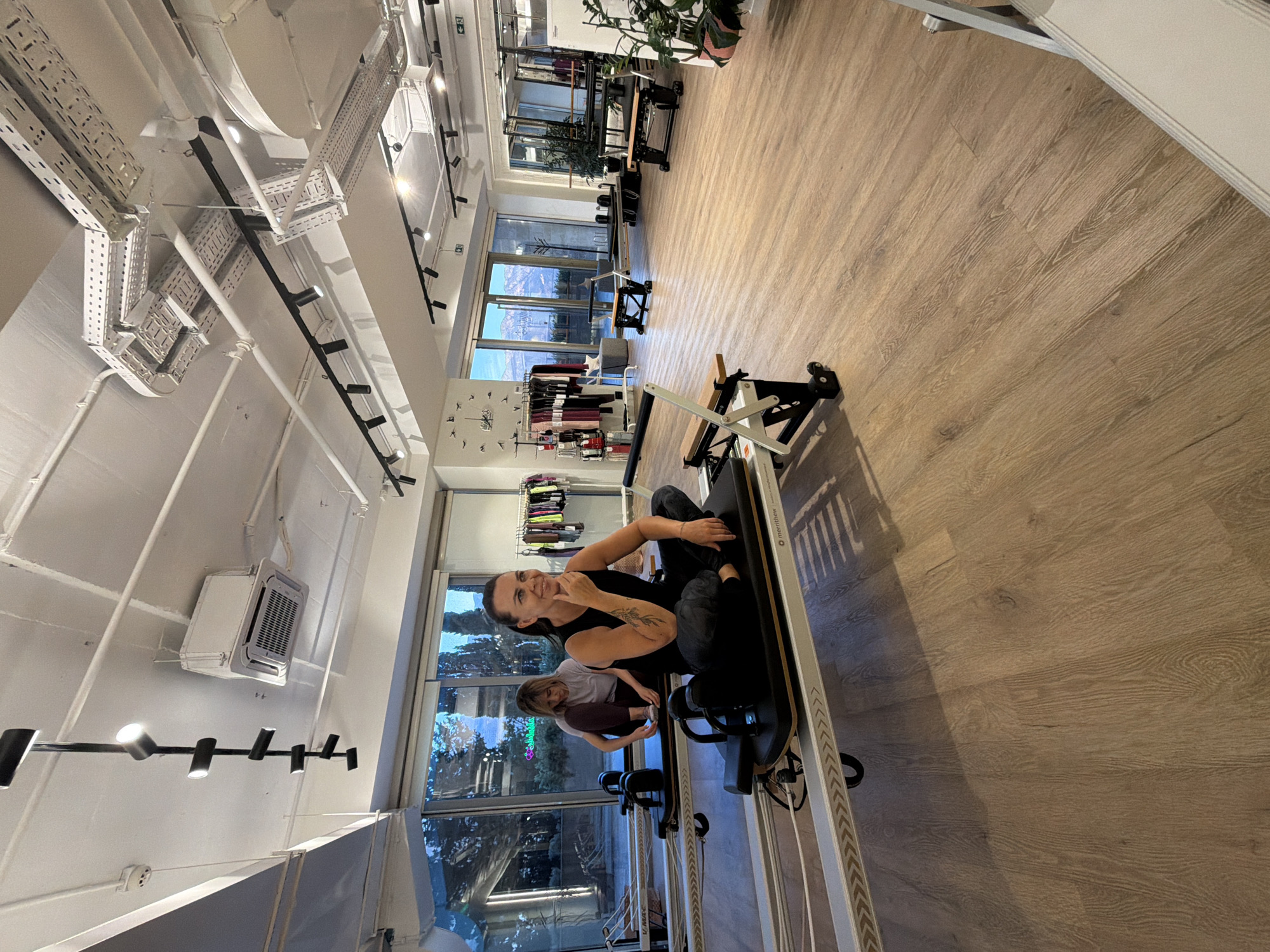 This is a medium-shot, eye-level, indoor photograph of a Pilates studio. In the foreground, a woman with dark hair tied back in a ponytail is sitting cross-legged on a Pilates reformer machine. She is wearing a black sleeveless top and dark patterned leggings. Her right hand is resting on her chin and she is looking to her right with a slight smile. Behind her, another woman with blonde hair is sitting on a similar Pilates reformer machine, facing away from the camera. She is wearing a white t-shirt and dark leggings and is reaching down towards her feet.
The studio has a light wood-look floor and a white ceiling with exposed industrial-style ductwork and lighting fixtures. Large windows in the background offer a view of a balcony and a mountainous landscape with trees. In the studio, there are several other Pilates reformer machines. To the left, there is a rack of athletic wear displayed. The overall atmosphere is clean and modern. The image was taken in Boreti, Montenegro.
