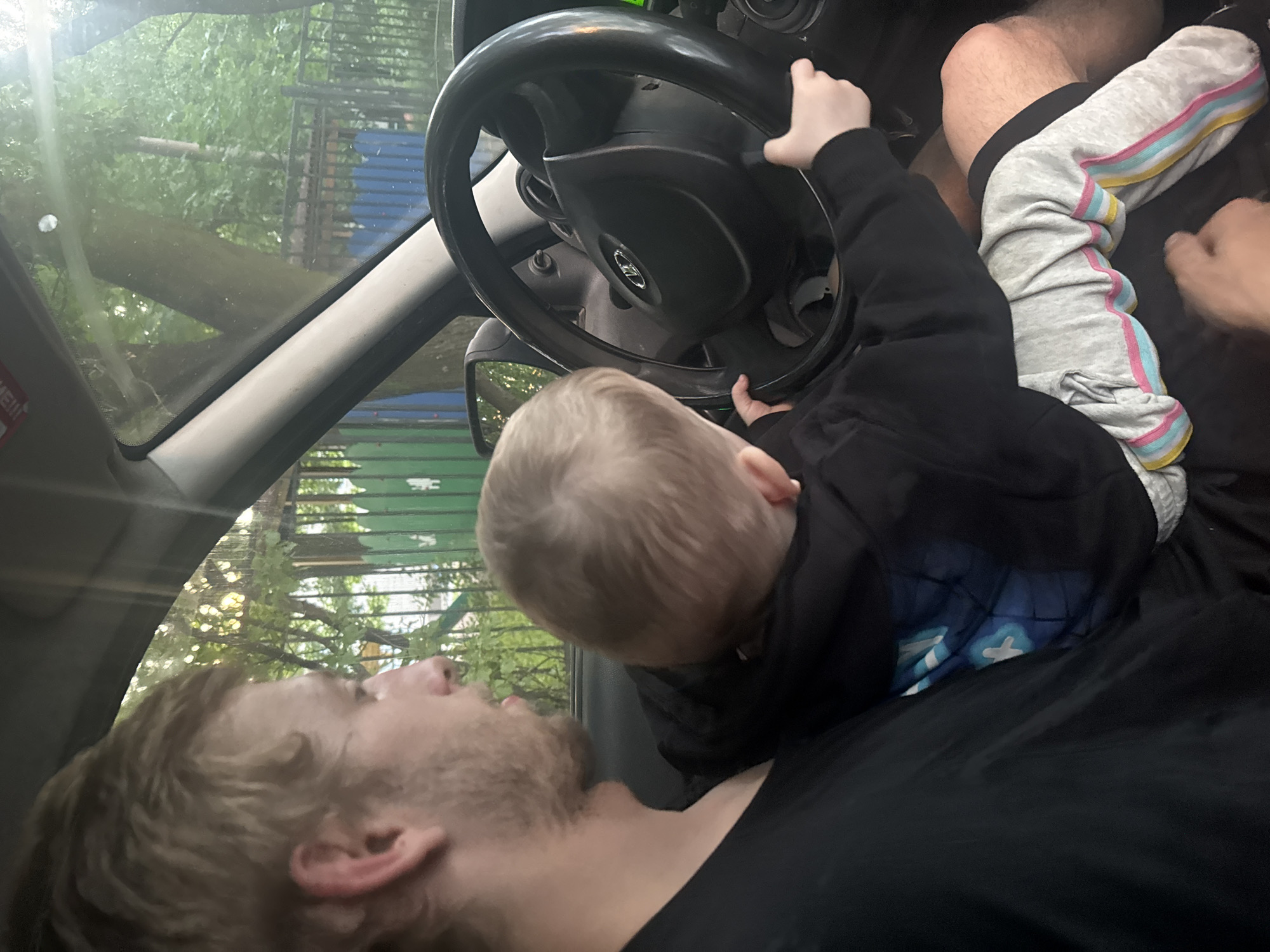 A man, with blond hair and a beard, is sitting in the driver's seat of a car, holding a young child on his lap. The child, with blond hair, is facing away from the camera and appears to be reaching for the steering wheel. The car's interior is visible, including the steering wheel with a logo that appears to be a Lada. Outside the car, through the windows, trees and a green fence are visible, suggesting an outdoor, possibly residential, setting. The lighting indicates it is daytime. The scene appears to be in Saint Petersburg, Russia, judging by the car model and general surroundings.