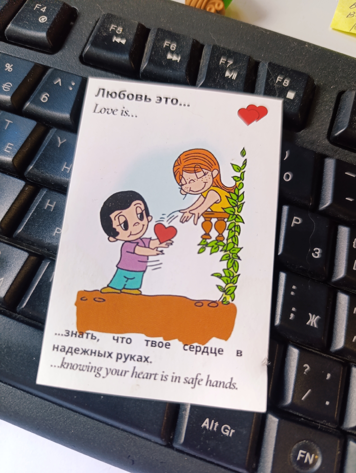 A small card is placed on a black computer keyboard. The card features cartoon illustrations of a boy and a girl with the text "Любовь это..." and "Love is..." at the top. The illustration depicts the boy offering a red heart to the girl, who is standing behind a fence covered in green leaves. At the bottom of the card is the text "...знать, что твое сердце в надежных руках." and "...knowing your heart is in safe hands." The keyboard shows keys such as F4, F5, F6, F7, F8, %, €, 6, T, Y, E, H, P, 3, :, Ж, ?, /, Alt Gr, and FN.