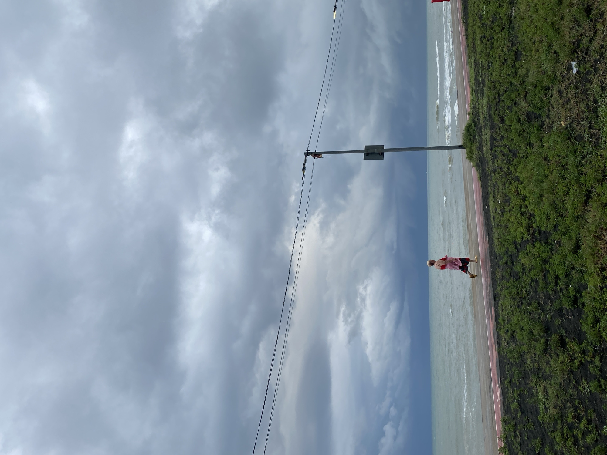 A lone individual, appearing to be an elderly woman with blonde hair, is depicted jogging along a red-lined promenade beside the sea. She is wearing a pink tank top and striped shorts. The sky above is heavily overcast with dark, dramatic clouds, suggesting impending stormy weather. The sea exhibits choppy waves, and a utility pole with a light fixture stands on the promenade. In the foreground, a patch of dark soil is covered with green grass. The scene is identified as being on the Ureki promenade in Ureki, Georgia.
