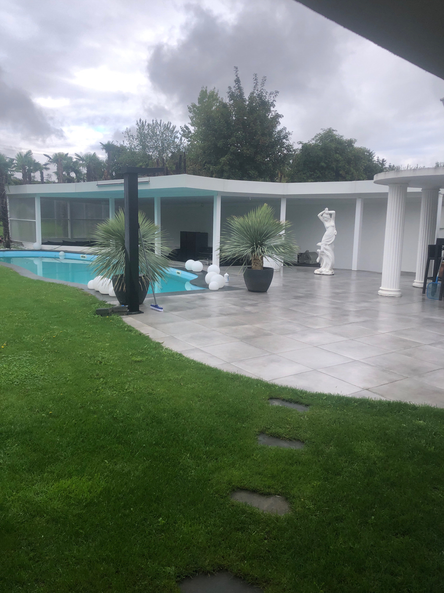 An outdoor paved patio with a swimming pool to the left, a lawn to the foreground, and a covered structure with white columns in the background. The sky is overcast. In the distance, palm trees are visible in front of the covered structure. A statue of a woman is present on the patio, and a few large white spherical objects are scattered around the pool area. A broom is leaning against a black pole near the pool. The scene appears to be a residential property located in Sierndorf, Austria.