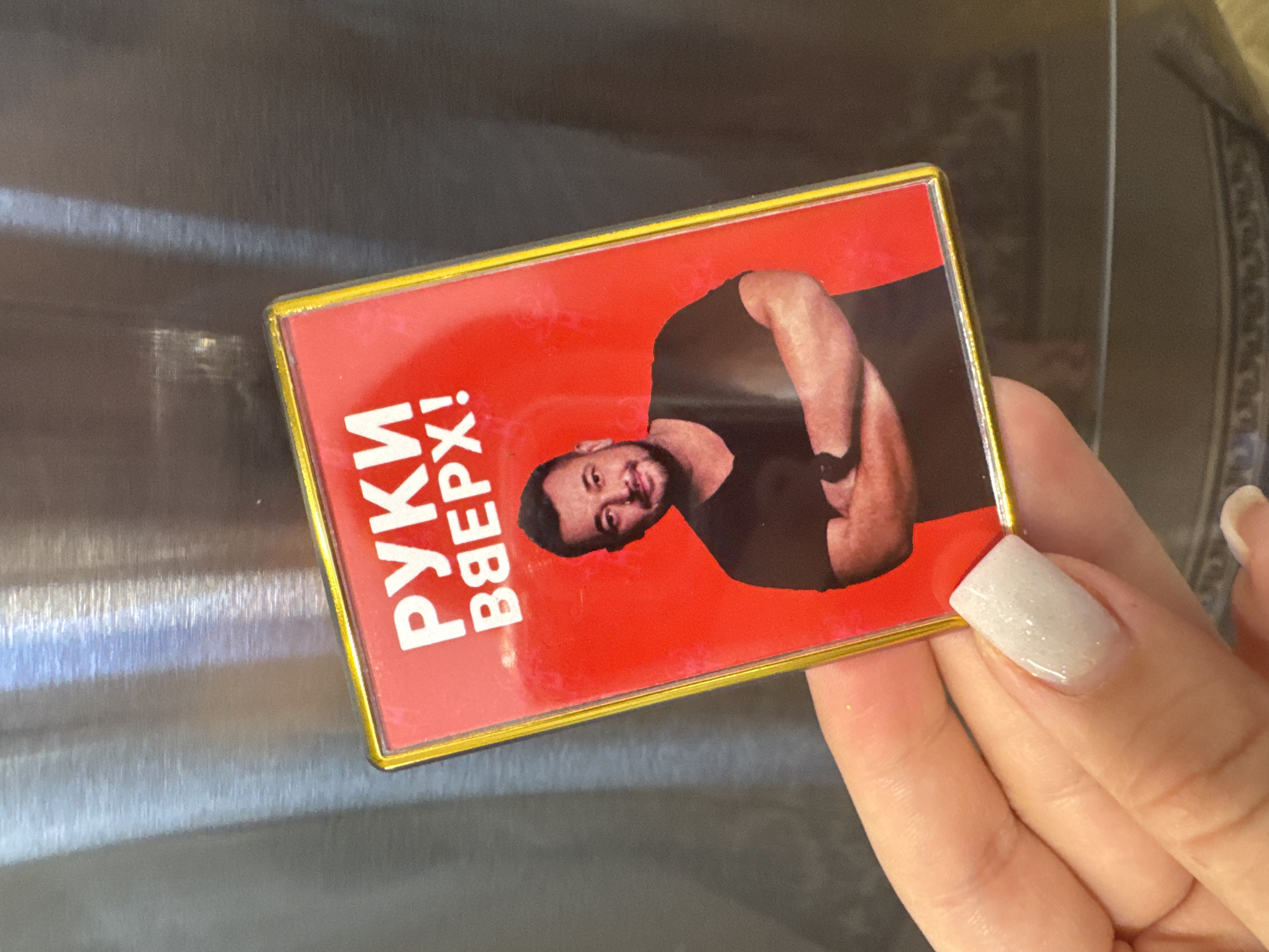 A rectangular magnet with a gold frame is being held by a fair-skinned hand with a glittery white nail. The magnet has a red background with the words "Руки Вверх!" written in white, vertical letters on the left. To the right is a photo of a man with a beard, wearing a black shirt, and his arms crossed. The magnet appears to be attached to a stainless steel surface. There is no discernible information to indicate the location as the city of Chaikovsky, Russia.