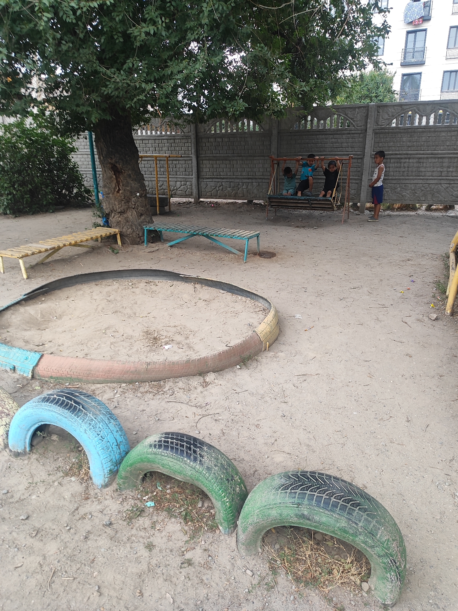 In an outdoor area, possibly a playground, several children are visible. Three boys are on a swing set, two are seated and one is standing. A fourth boy stands to the right of the swing set, facing away from it. In the foreground, large, colorful tires are arranged on the sandy ground. A sandbox with a tire border is situated behind the tires. A wooden bench is to the left of the sandbox, and another bench, painted blue, is to the right. A large tree with dense foliage dominates the left side of the image, casting shadows. In the background, a concrete fence separates the play area from a modern building with balconies. The overall environment appears to be a somewhat worn but functional outdoor recreational space.