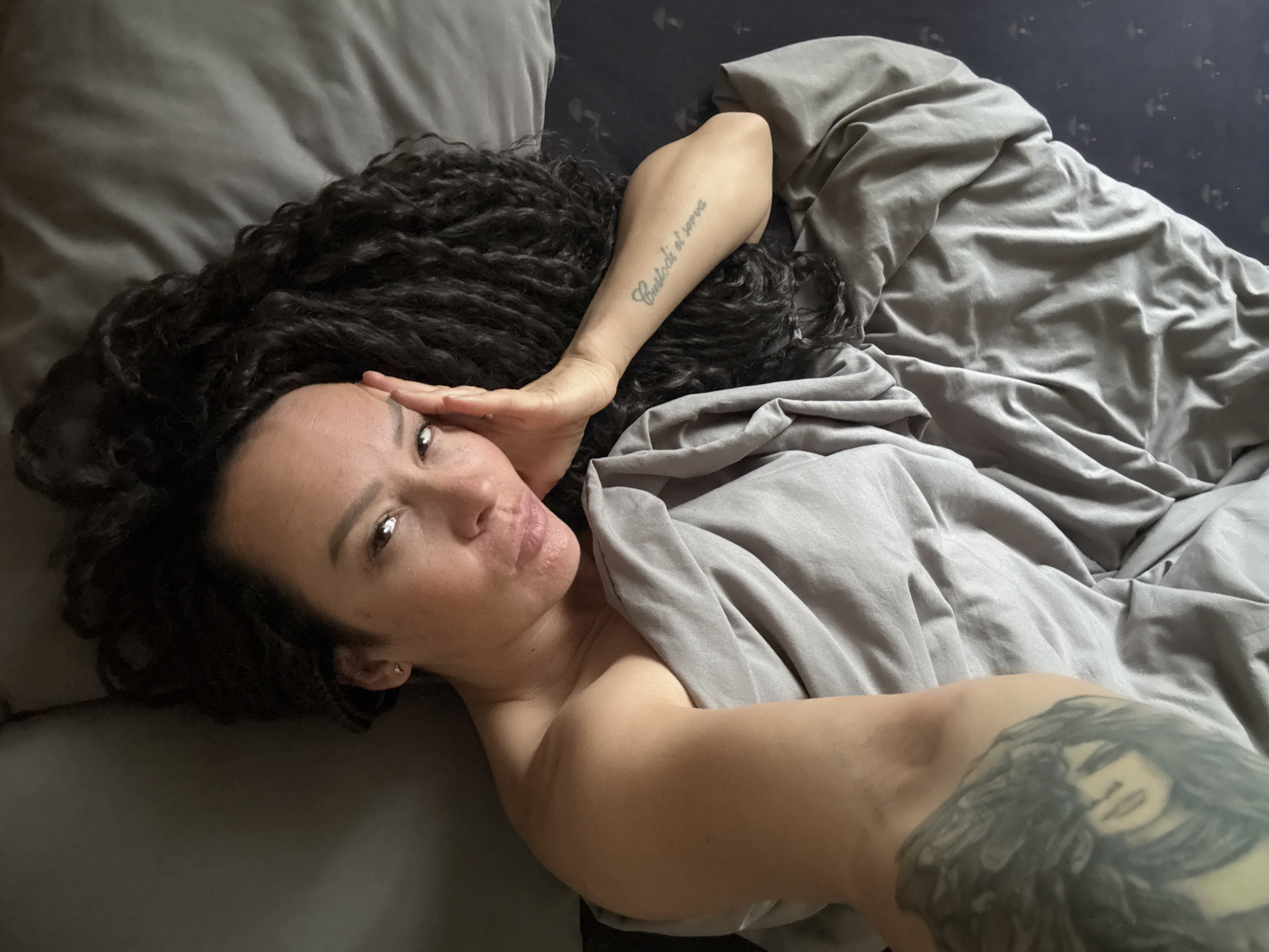 A woman is observed lying in a bed within an indoor residential setting in the city of Chaikovsky, Russia. The woman, appearing to be in an overhead selfie perspective, has long, dark, curly hair splayed around her head on a dark pillow or headboard. She looks directly at the camera with a slight pout, her left hand positioned with fingers resting against her left temple and cheek. Her right earlobe contains a small, light-colored stud earring. Her upper torso is partially covered by a light grey crumpled sheet. Her bare right arm displays a tattoo on the forearm reading 'Custodi et serva'. Her bare left upper arm features a large grayscale tattoo depicting a stylized face looking upwards. The fitted sheet beneath her is dark blue with a repeating pattern of light-colored, small, mushroom-like figures.