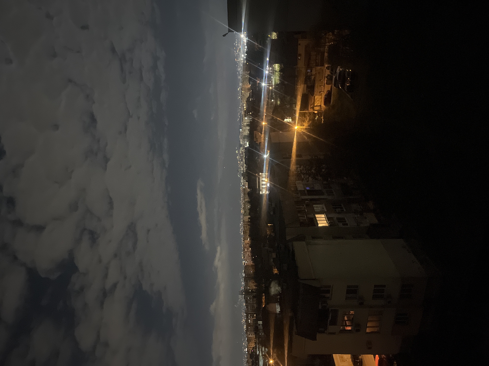 The image depicts a night cityscape. On the left, the sky is filled with heavy, dark clouds. In the middle, a line of lights stretches across the horizon, indicating a city skyline. On the right, buildings stand in the foreground, with a few windows lit up.
The location is likely Sofia, Bulgaria, based on the urban density, building styles, and general atmosphere. The scene captures a view of the city at night, with the skyline illuminated against the backdrop of a cloudy sky.