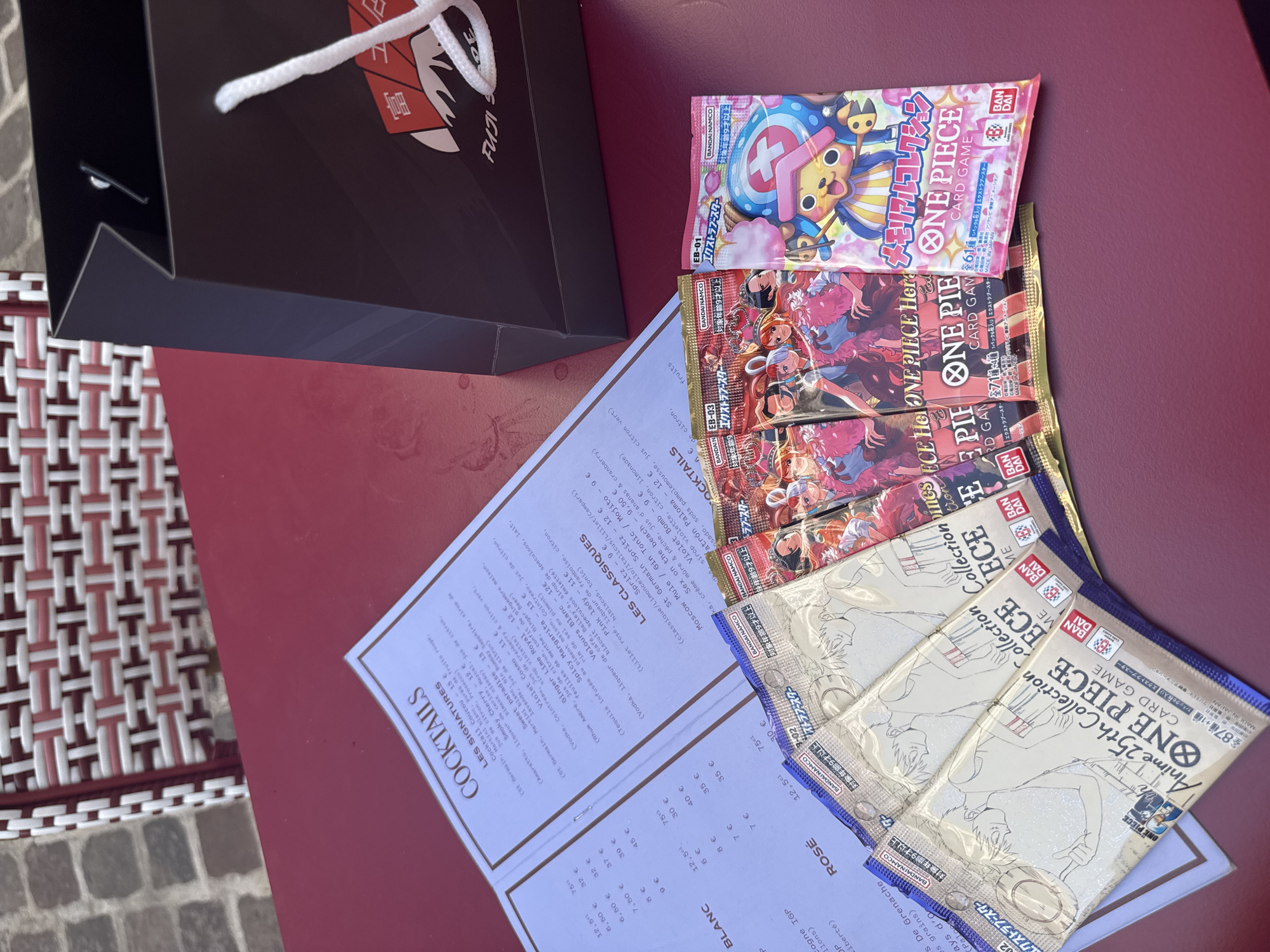 The image depicts a table with a red surface, likely outdoors due to the checkered pattern of a chair visible in the background. On the table, a cocktail menu, a black shopping bag with the Fuji logo, and several packs of "ONE PIECE" collectible card games are arranged. The menu displays cocktail names and prices. The shopping bag appears to be from a store, possibly a souvenir shop or a retailer. The card game packs are colorful and feature characters from the "One Piece" anime and manga series. There are no people visible in the image, and no actions are being performed. The scene suggests a casual setting, perhaps a cafe or restaurant, where someone has purchased these items. The mention of Lille, France, in relation to this image is not supported by any visual cues within the image itself.
