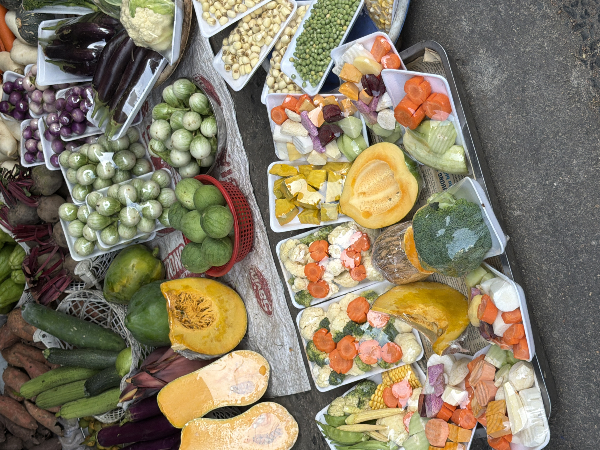 An overhead, eye-level medium shot displays a variety of fresh produce at a street market in Nha Trang Ward, Vietnam. The scene is dominated by an assortment of vegetables and fruits, many of which are pre-cut and packaged in plastic trays. In the foreground, several trays contain an array of chopped vegetables, including carrots, broccoli, cauliflower, and pieces of squash and possibly sweet potato. A large, halved yellow squash with visible seeds sits prominently in the lower center. To the left, two halved butternut squash are displayed. Moving towards the background, trays of smaller, round green vegetables resembling green peas are visible, as well as lotus seeds. Other vegetables such as eggplants, zucchini, and what appear to be small, round, light-green eggplants are arranged in baskets and trays. The produce is laid out on a textured surface, possibly a tarp or cloth, with some newspaper visible underneath. The overall impression is a vibrant and colorful display of fresh food for sale.