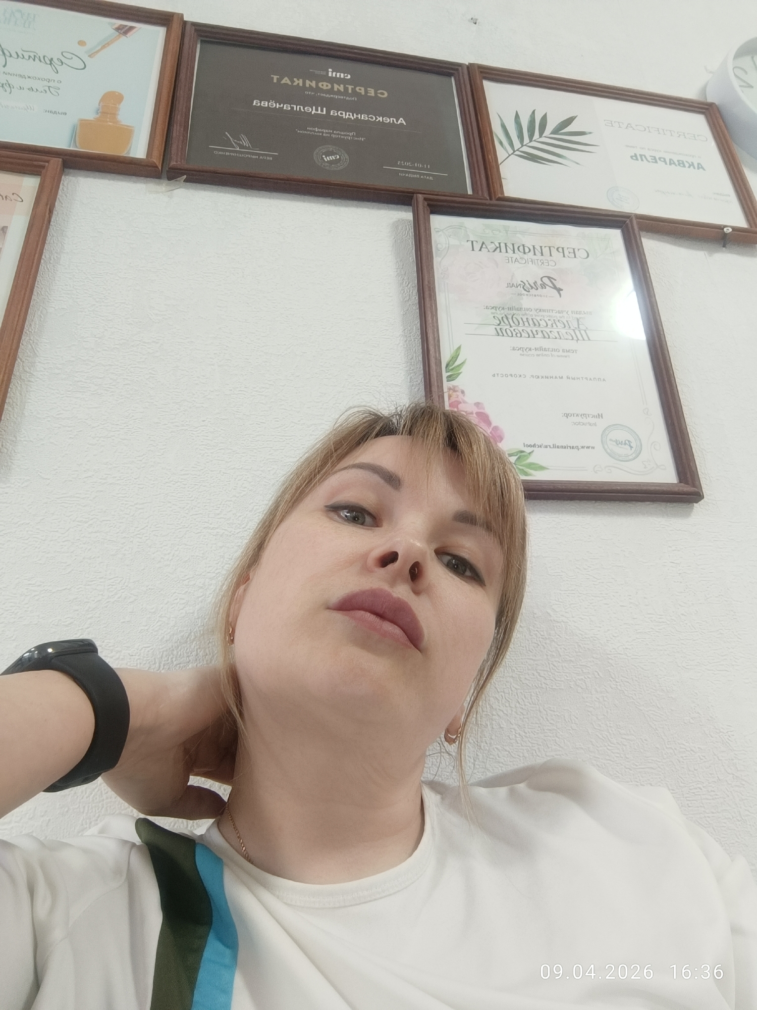 Here is a forensic description of the image:
**Scene:** A selfie taken indoors against a textured, light-colored wall decorated with several framed certificates and documents. The picture is angled upward, focusing on a woman.
**Subject:** The primary subject is a fair-skinned woman with blonde hair and light eyes. She wears a white T-shirt with green and blue stripes on the shoulder and a black fitness tracker watch on her left wrist. She has a neutral expression.
**Objects:**
* Framed certificates and documents hanging on the wall behind the woman. One certificate is visible behind her head and has floral decorations and is likely a training or qualification certificate. The certificates are of varying sizes and styles, with wooden frames.
* Black fitness tracker.
* Gold earrings.
**Actions/Interactions:** The woman is taking a selfie. Her head is tilted back slightly.
**Environment:** The location appears to be an office or business setting, judging by the certificates displayed.
The time stamp is displayed at the bottom right corner: 09.04.2026 16:36.
I am unable to find the location in the city of Chaikovsky, Russia.