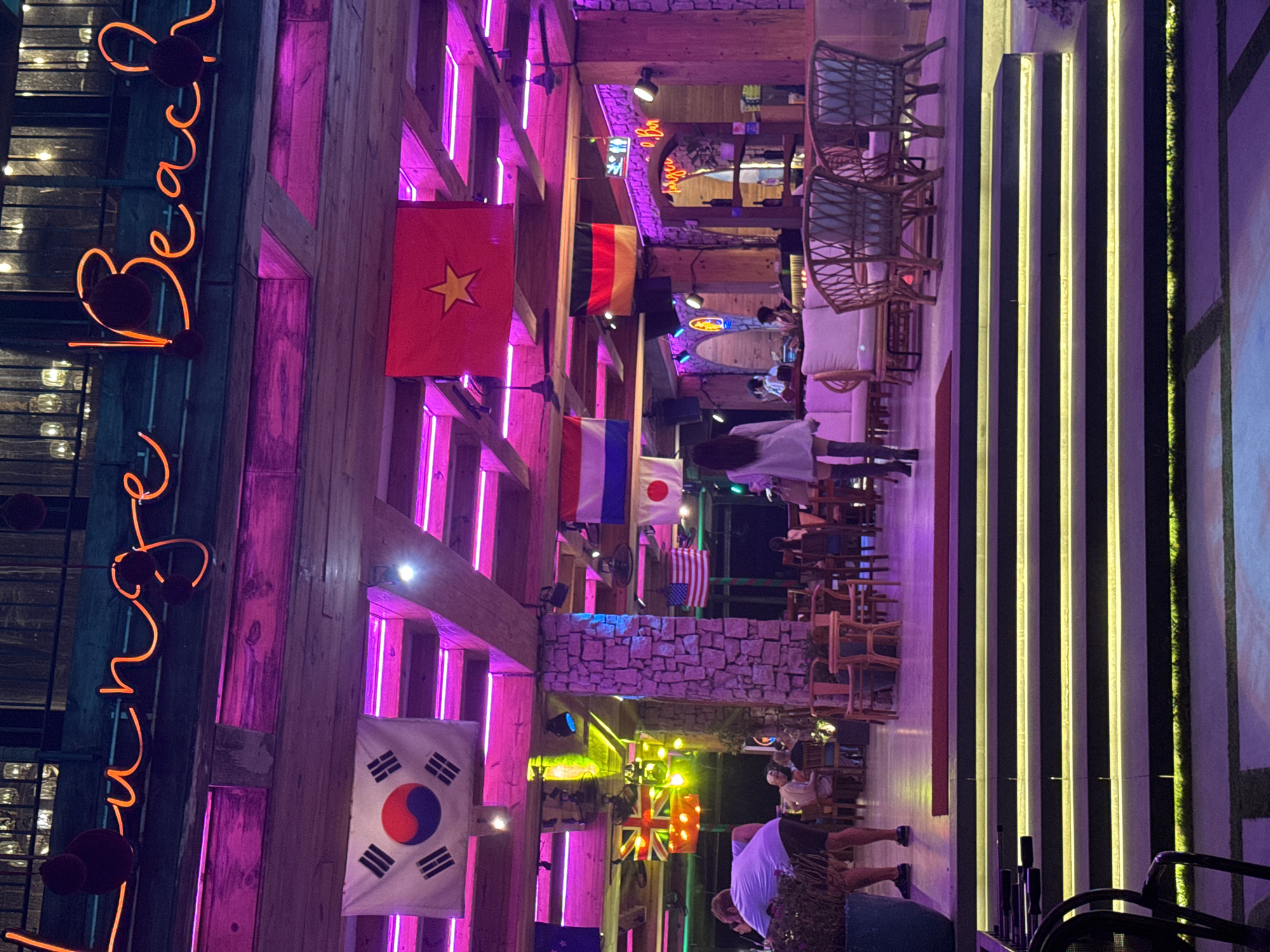 The image shows an indoor area in what appears to be a beach club or restaurant, illuminated with neon lights and decorated with flags of various nations. The text "Sunrise Beach" is visible in neon signage. The building has wooden beams and stone accents, and the overall lighting is a mix of pink, purple, and yellow neon. There are tables and chairs set up, and people are present. The location is likely in Đà Nẵng, Vietnam.